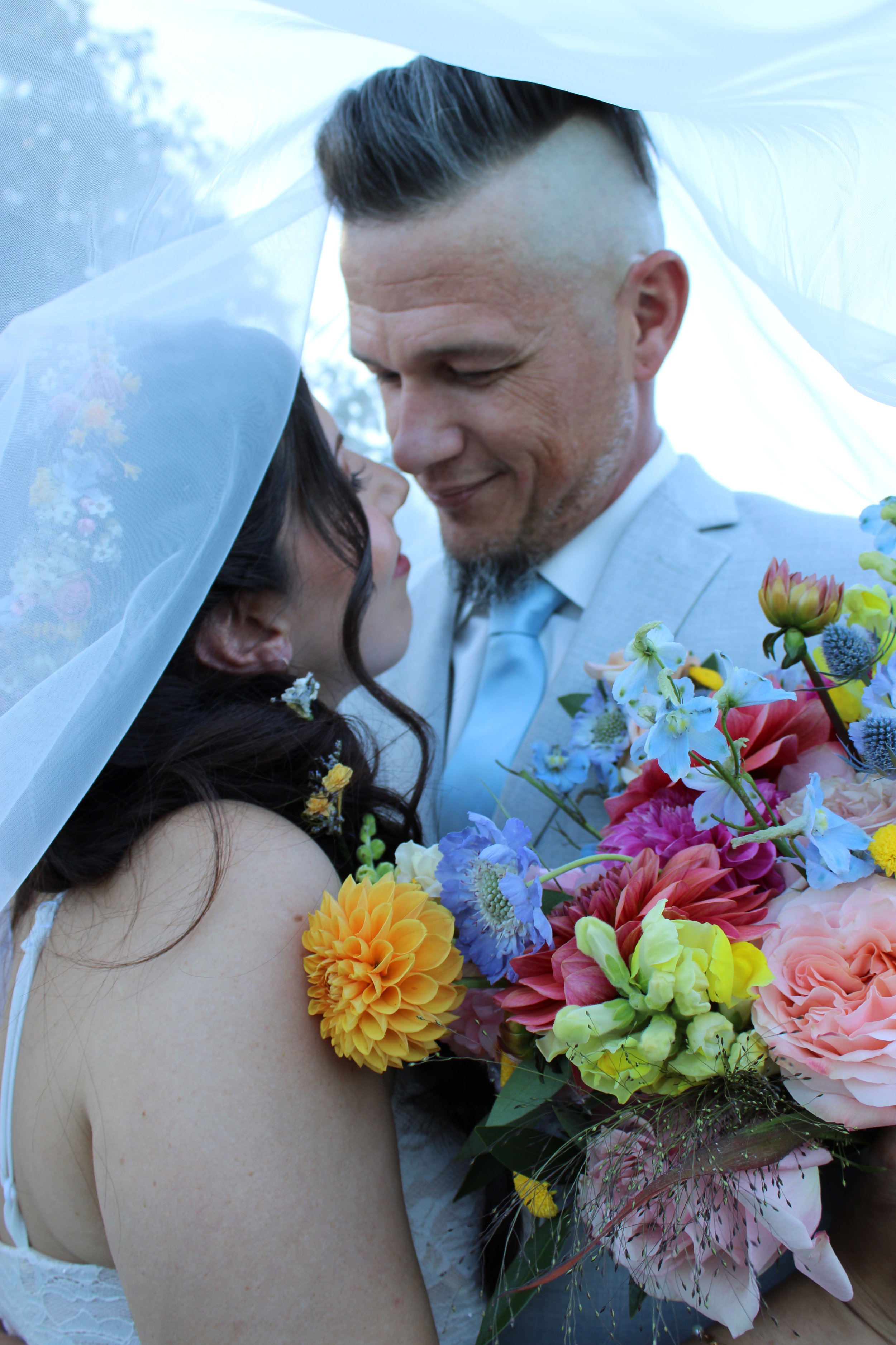 A man and woman in wedding attire embrace, with the woman holding a large bouquet of colorful flowers, including yellow, pink, orange, and blue blossoms, under a sheer white veil.