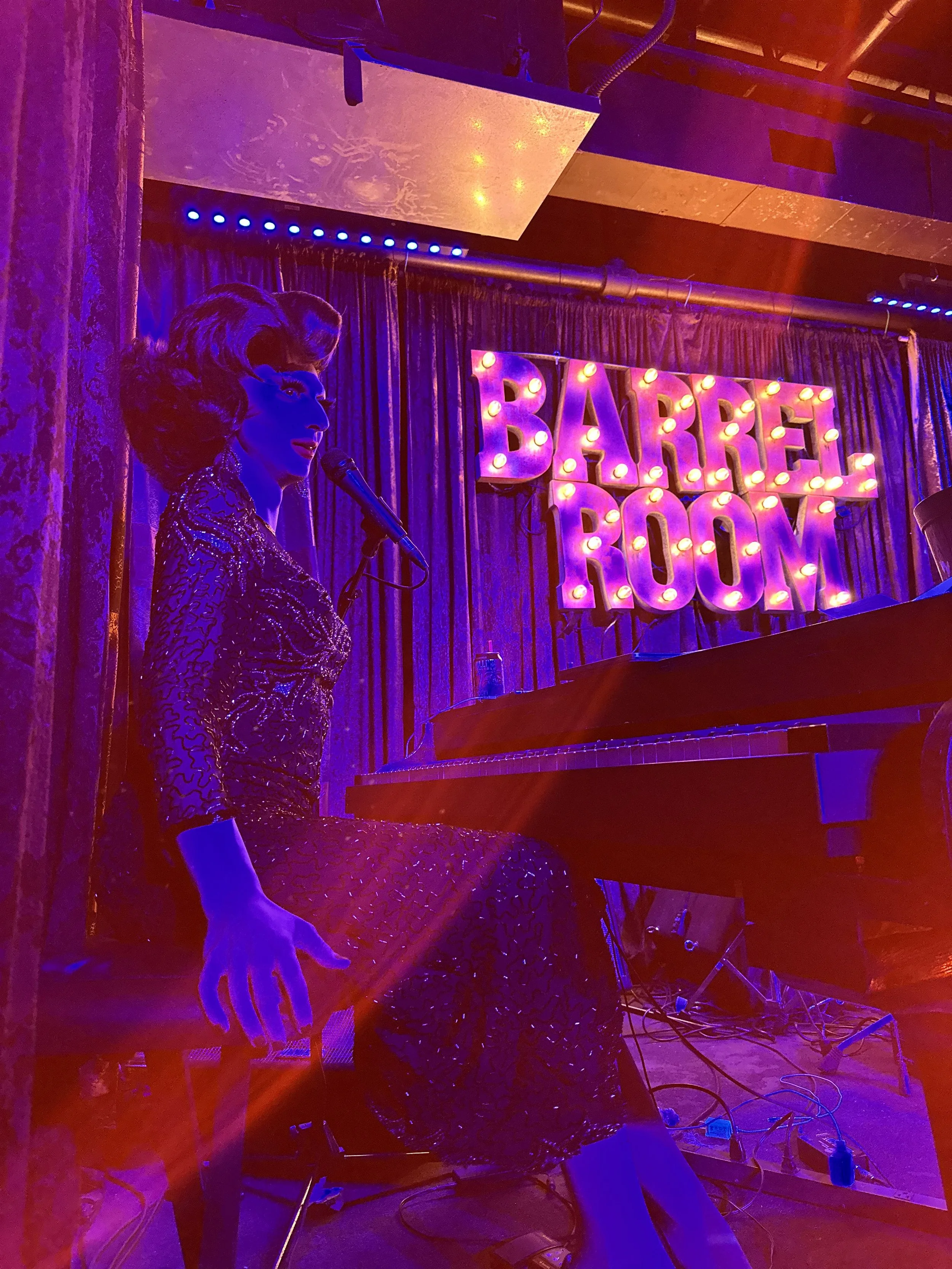 A drag queen performing on stage with a microphone, dressed in a patterned dress, illuminated by purple and pink stage lights, with a sign reading 'Barrel Room' in the background.