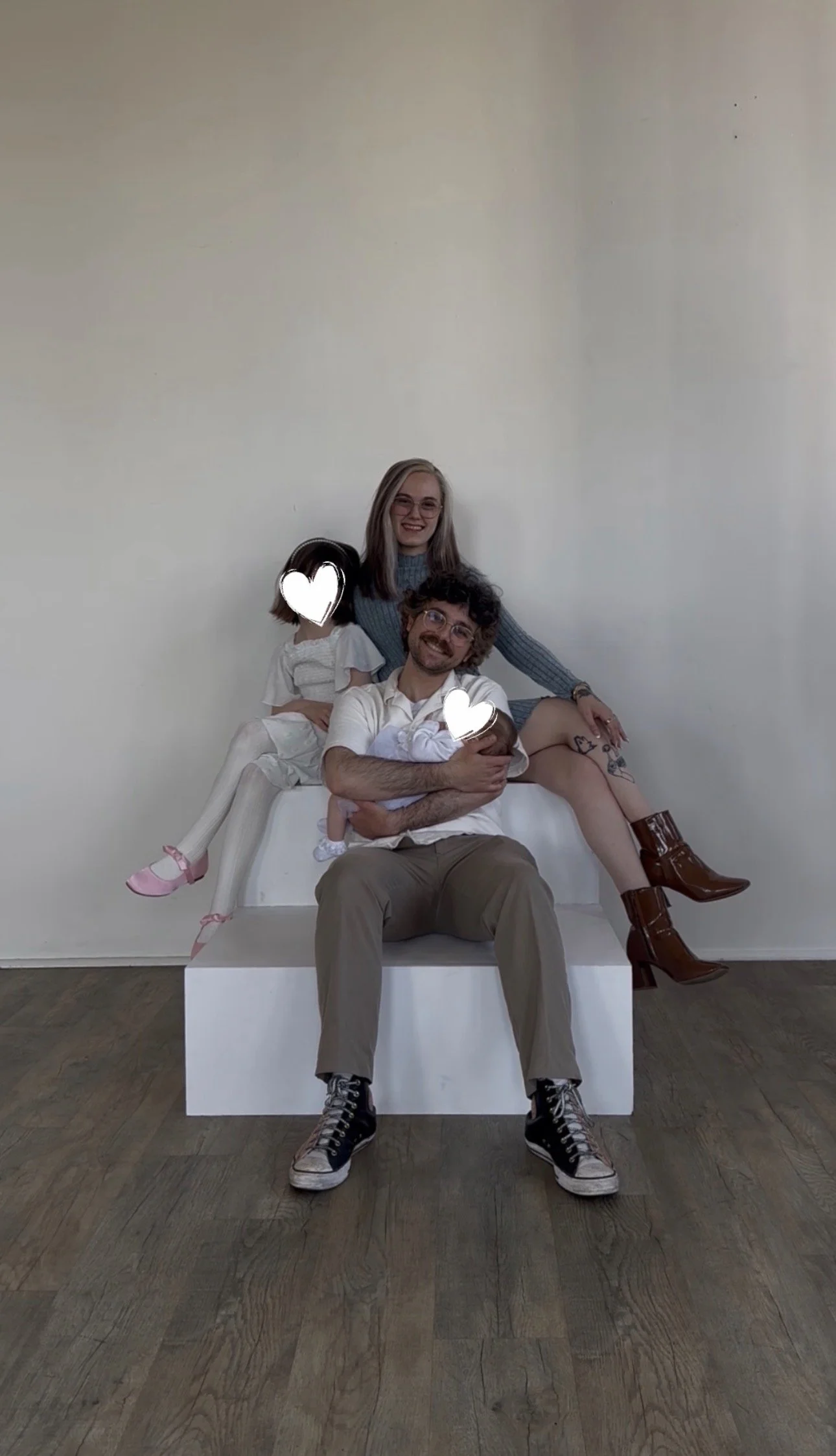 A family of four sitting on a white bench against a plain wall. The father is in the center with a small baby in his arms, while a woman and a girl are sitting on either side of him. The woman's face is visible, and she is smiling. The girl on the left has her face obscured with a heart shape. The woman is wearing a long-sleeved shirt, the girl is wearing a white dress, and the father is wearing a white shirt and gray pants. The woman is wearing brown ankle boots, and the girl is wearing pink shoes. The family appears happy and relaxed.