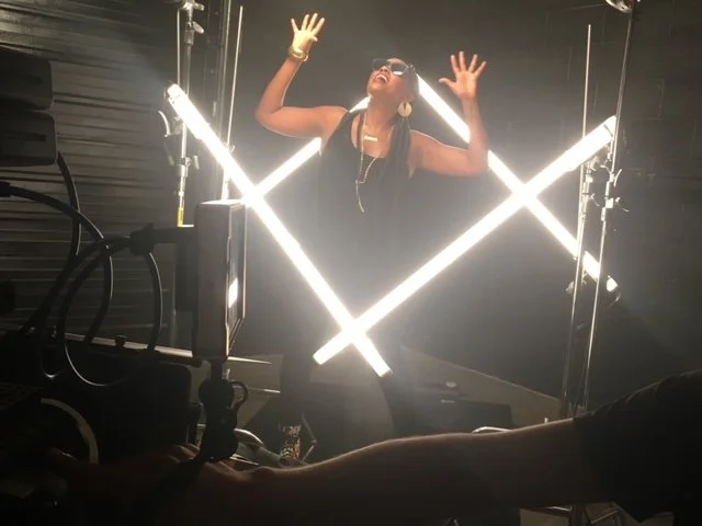A woman wearing sunglasses, headphones, and jewelry, standing behind luminous crossed light bars, singing or speaking with hands raised in a dark setting, possibly a recording studio or stage.