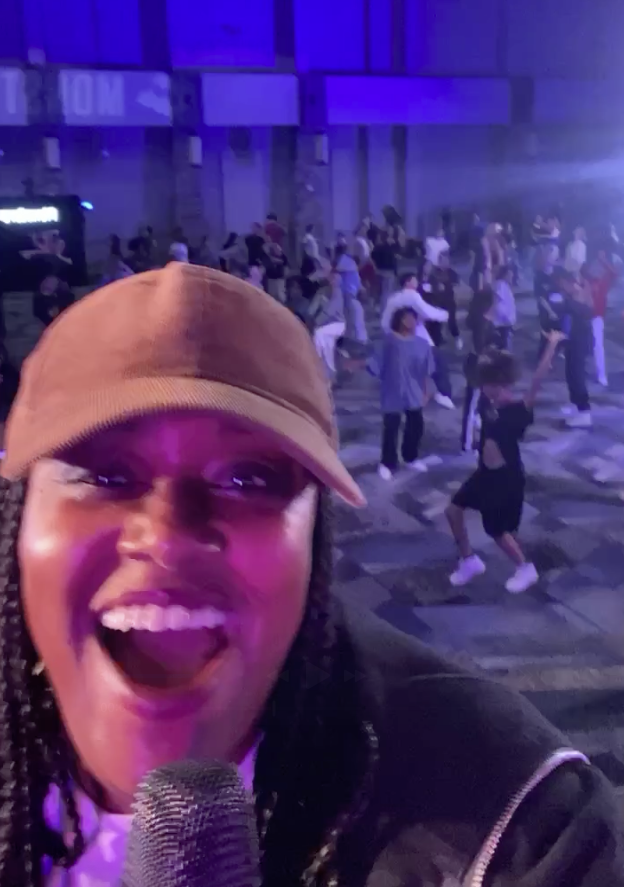 A woman smiling and singing into a microphone at a lively indoor event with many people dancing in the background, illuminated by colorful lights.