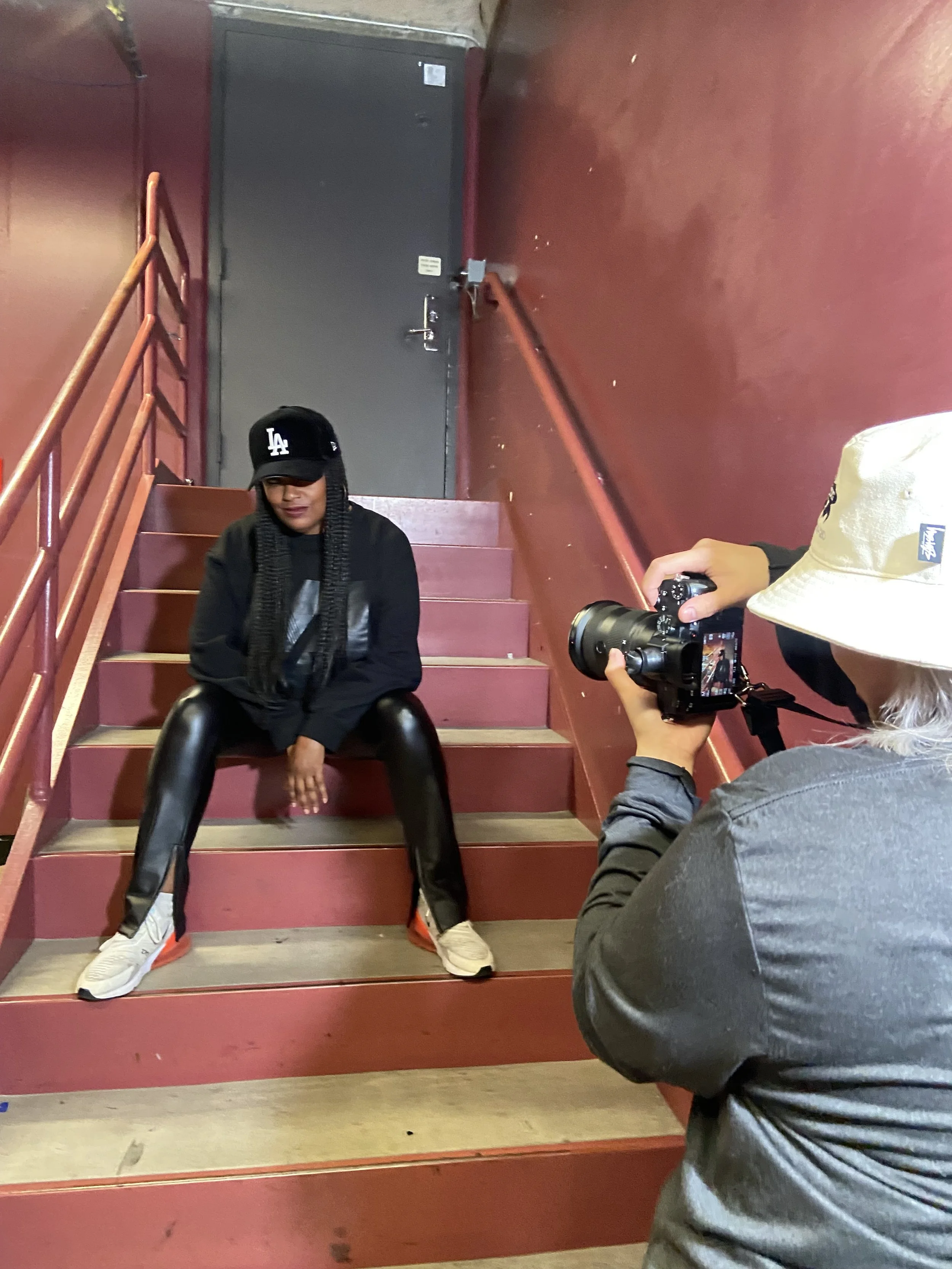 A woman sitting on red staircase with black leather pants, black sweatshirt, and a black baseball cap, posing for a photo being taken by a person with a camera. The setting appears to be indoors with red walls and a black door in the background.
