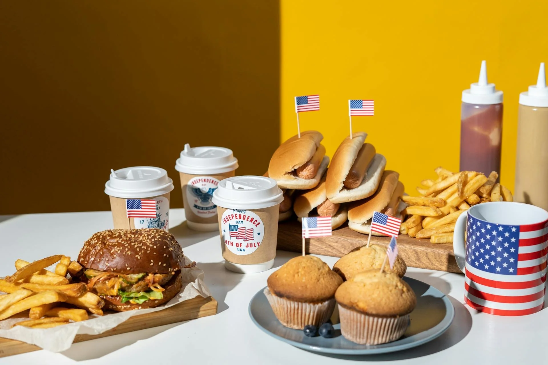 American-themed food and drinks for Independence Day, including burgers, hot dogs with small American flags, french fries, muffins, and coffee cups with patriotic designs, set against a yellow and brown background.