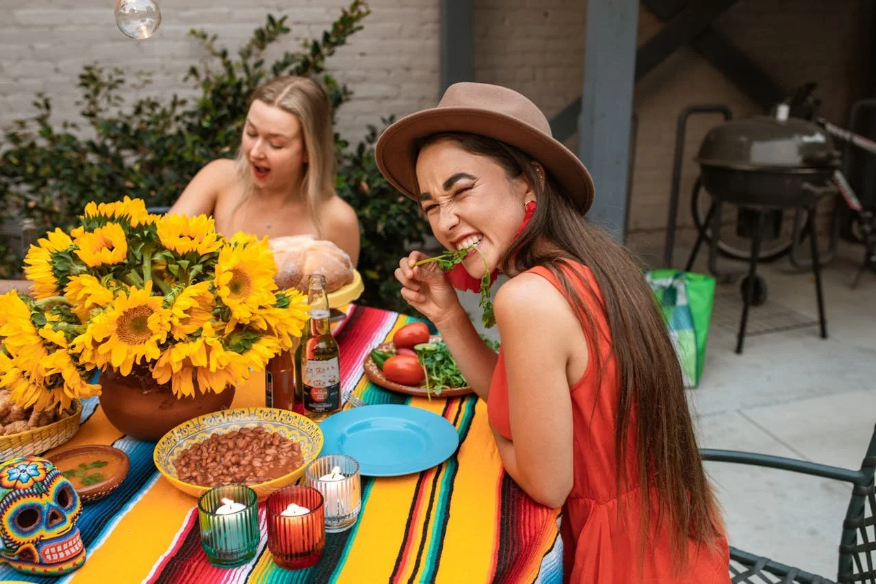 Two women enjoying a festive meal outdoors at a decorated table with sunflowers, candles, and traditional Mexican decorations; one woman is in focus, wearing a hat and a red dress, laughing and eating a leaf, while the other woman is in the backgroun