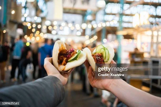 Two hands holding steamed buns with filling and cucumber slices at a food market