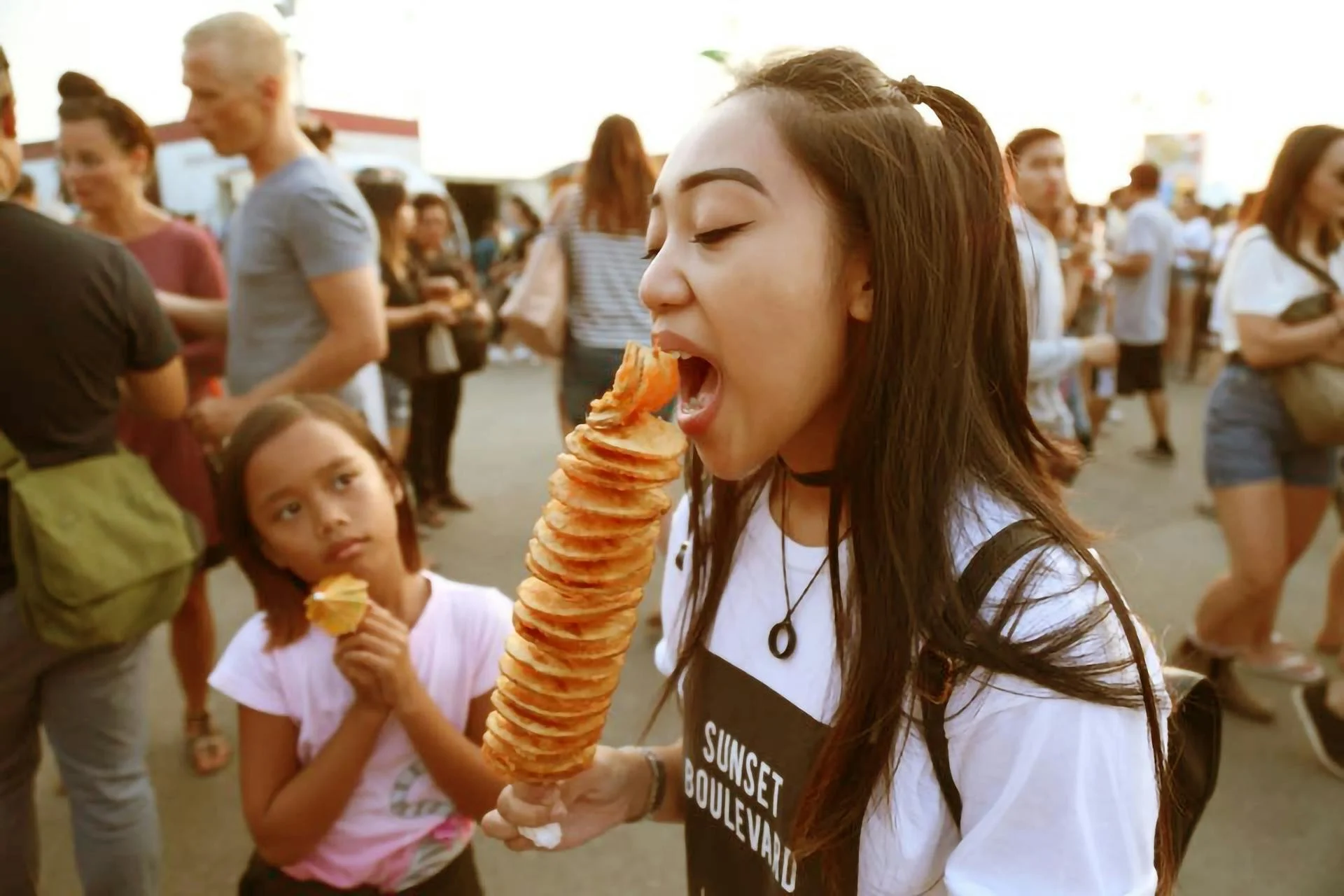 A young woman with dark hair is about to take a bite from a giant spiral potato on a stick at an outdoor fair, with a young girl in a pink shirt nearby holding a smaller snack, and a crowd of people in the background.