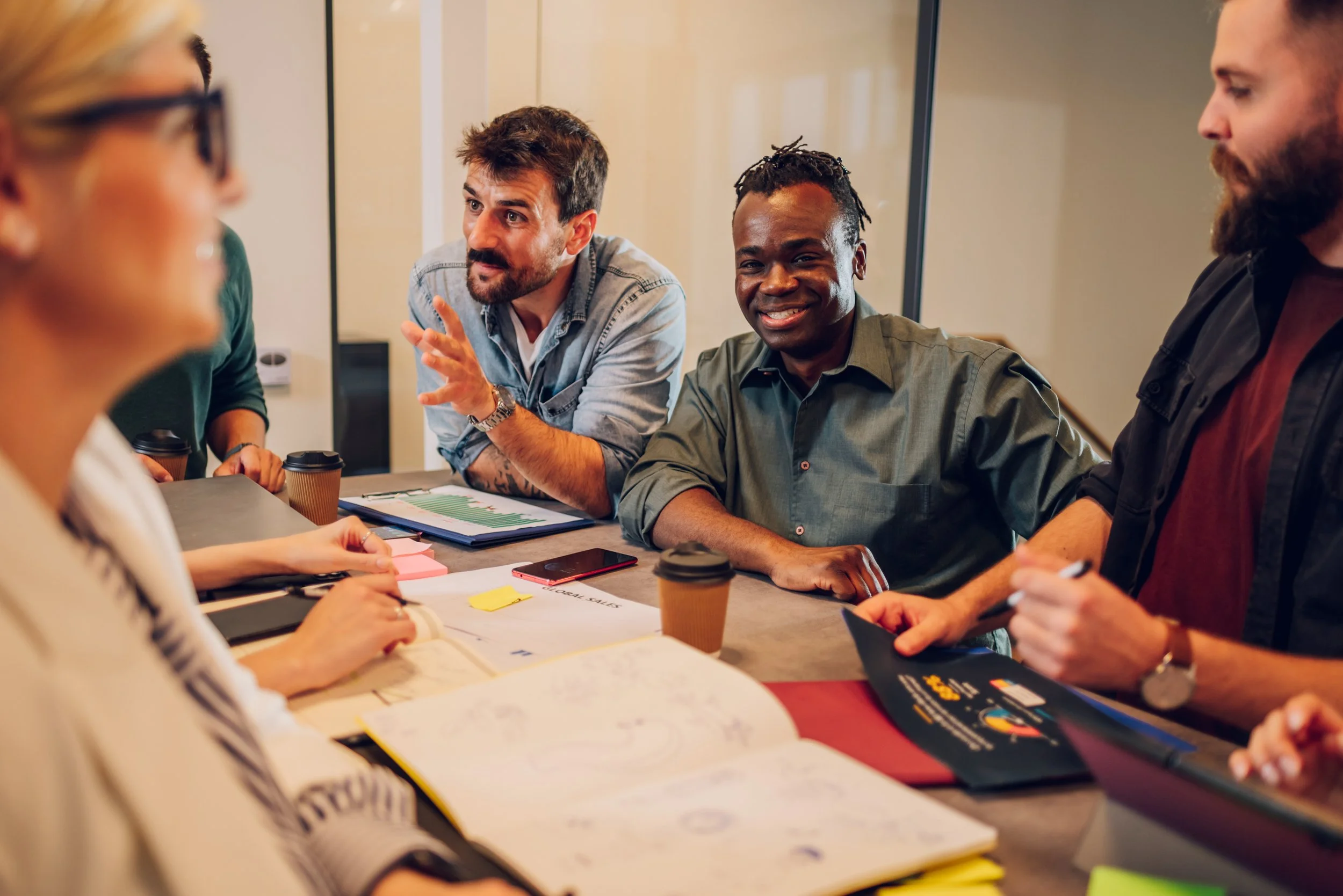 A diverse group of five people engaged in a discussion around a conference table with documents, notebooks, and coffee cups.