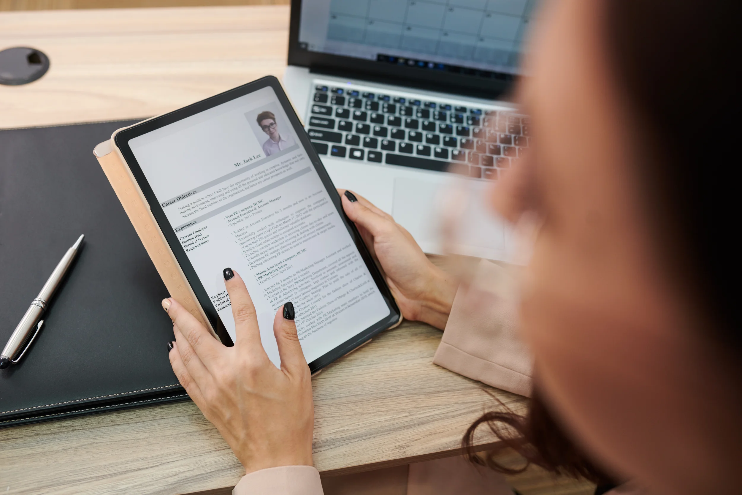 Person holding a tablet displaying a resume, with a laptop and pen on the desk.