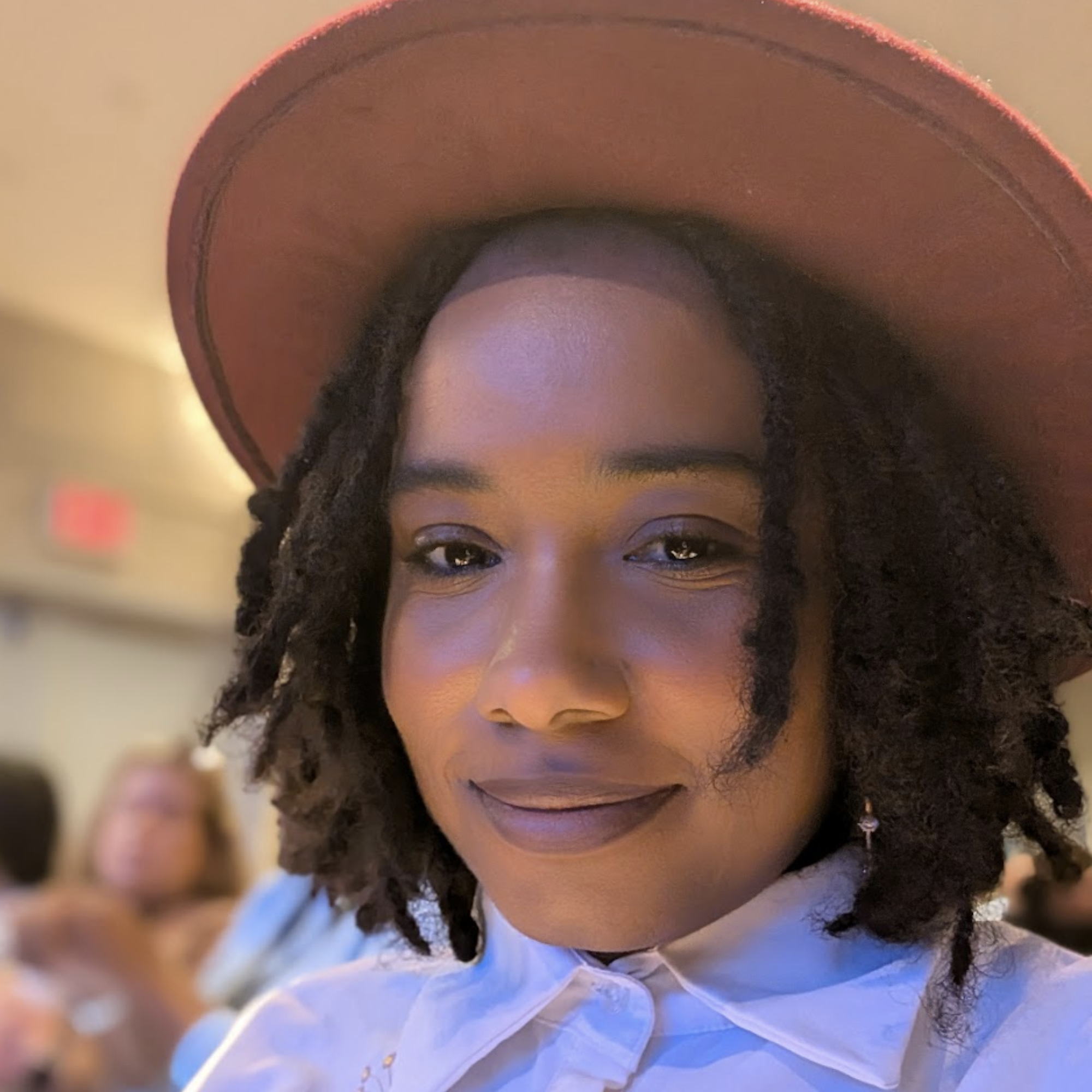 A woman with dark skin and natural curly hair wearing a large brown hat and a white collared shirt, smiling at the camera in an indoor setting.