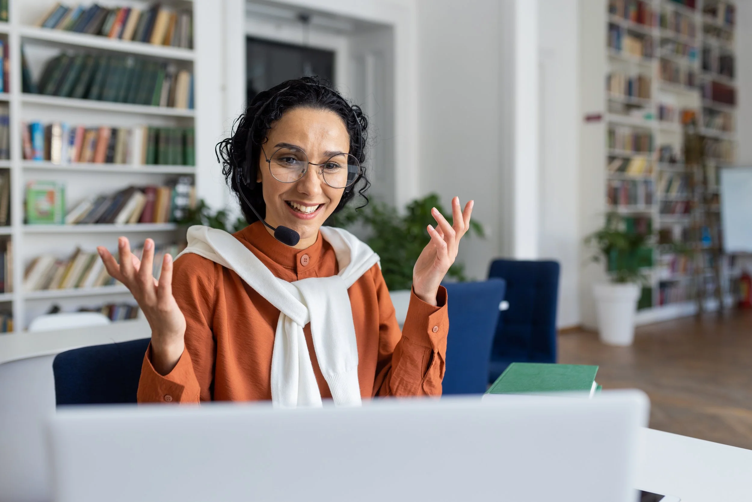 A woman wearing glasses and a headset sits at a desk, smiling and gesturing with her hands in a library or office with bookshelves and plants in the background.