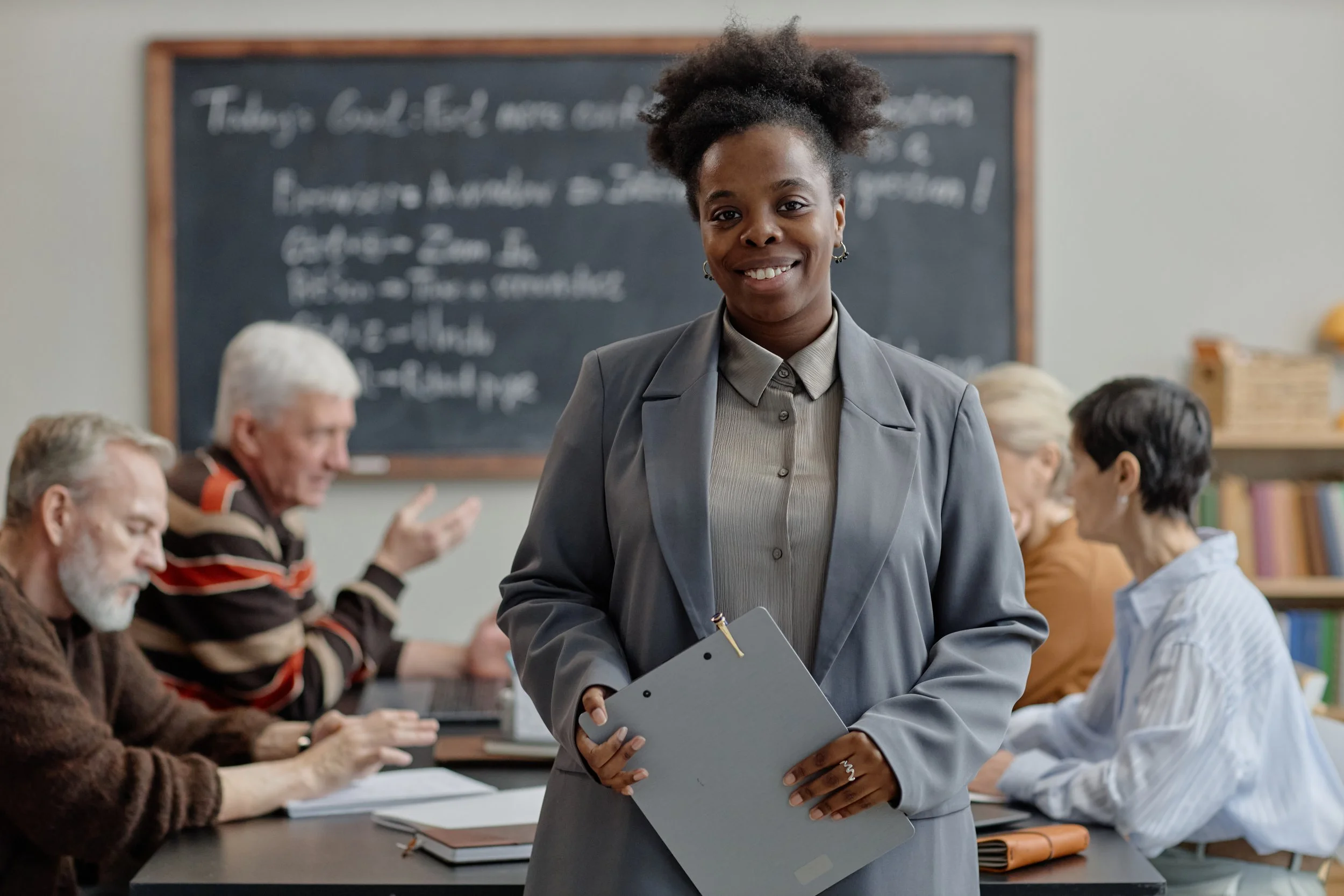 A professional woman standing in a classroom or conference room, holding a gray folder and smiling at the camera. In the background, several older adults are engaged in conversation, with a blackboard behind them.