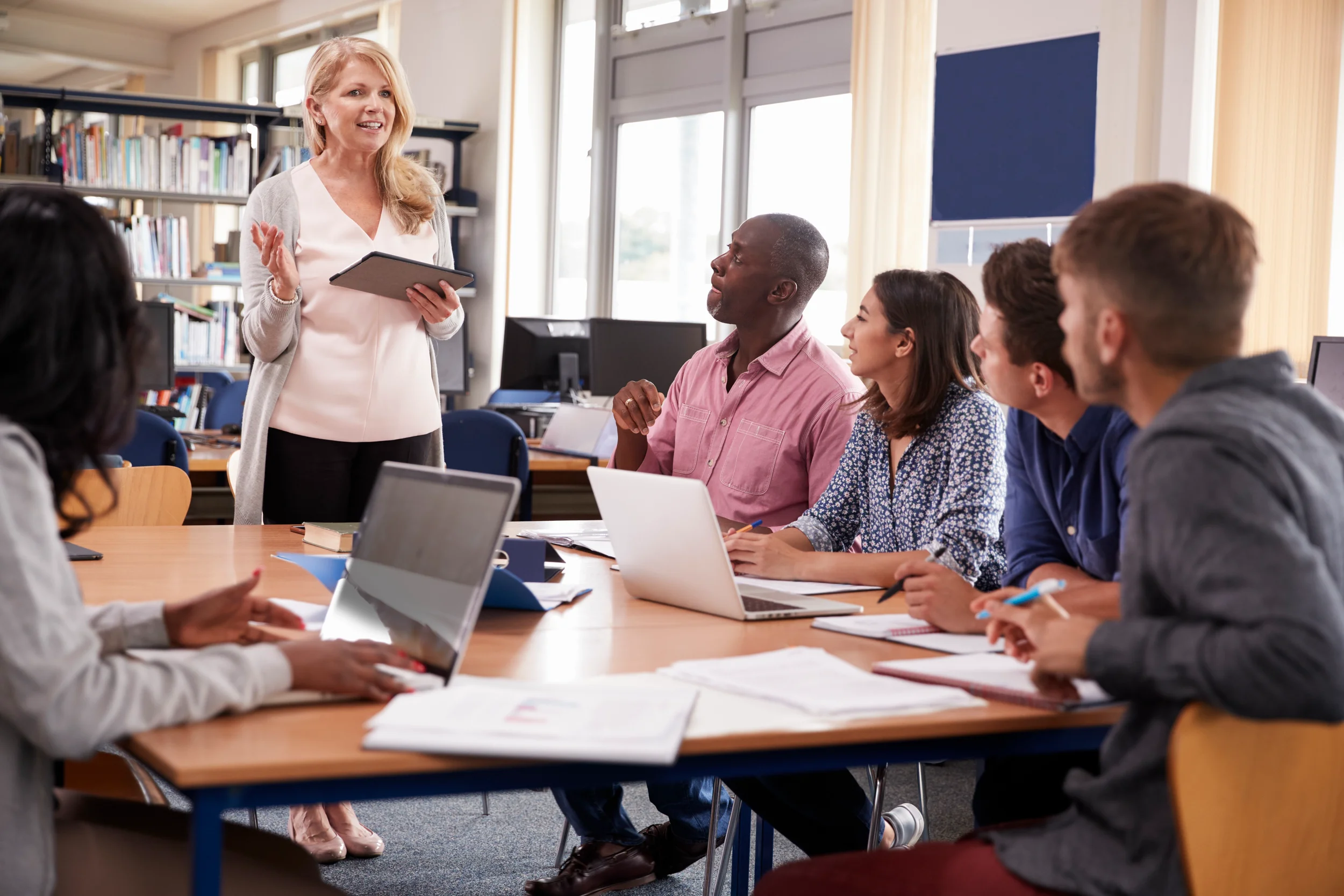 A woman standing and smiling while speaking in front of a group of diverse people sitting at a table with laptops and notebooks in a classroom or meeting room.