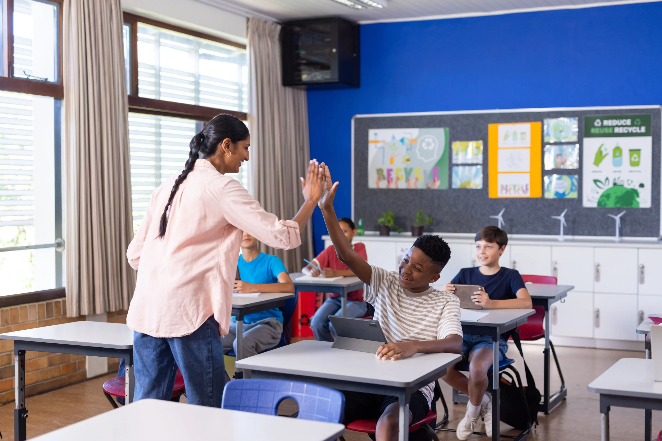 A classroom with a teacher giving a high-five to a student who is smiling, with other students seated at their desks and colorful environmental posters on the wall.