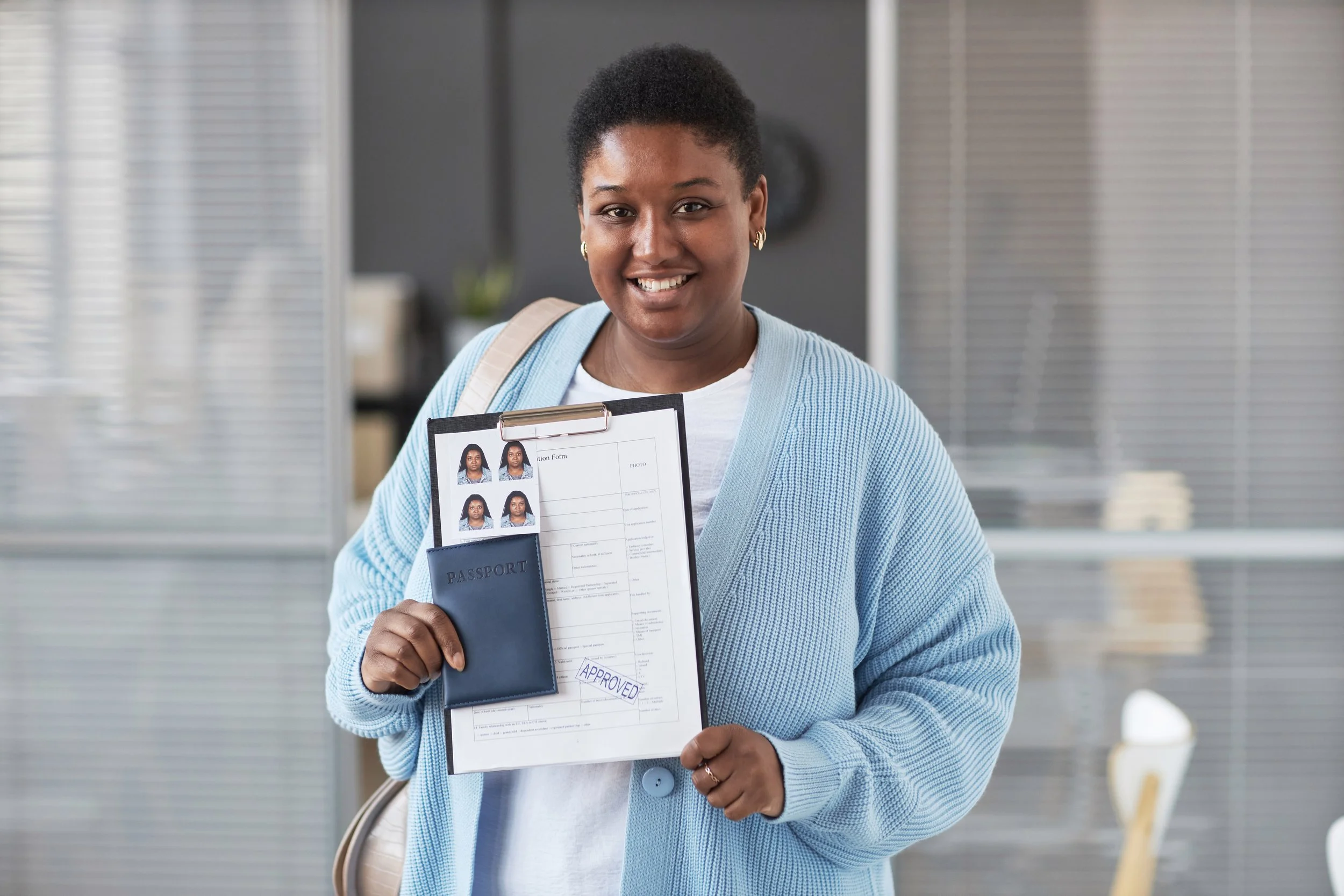 Smiling woman holding a clipboard with passport photos, a form, and an approved stamp in an office setting.