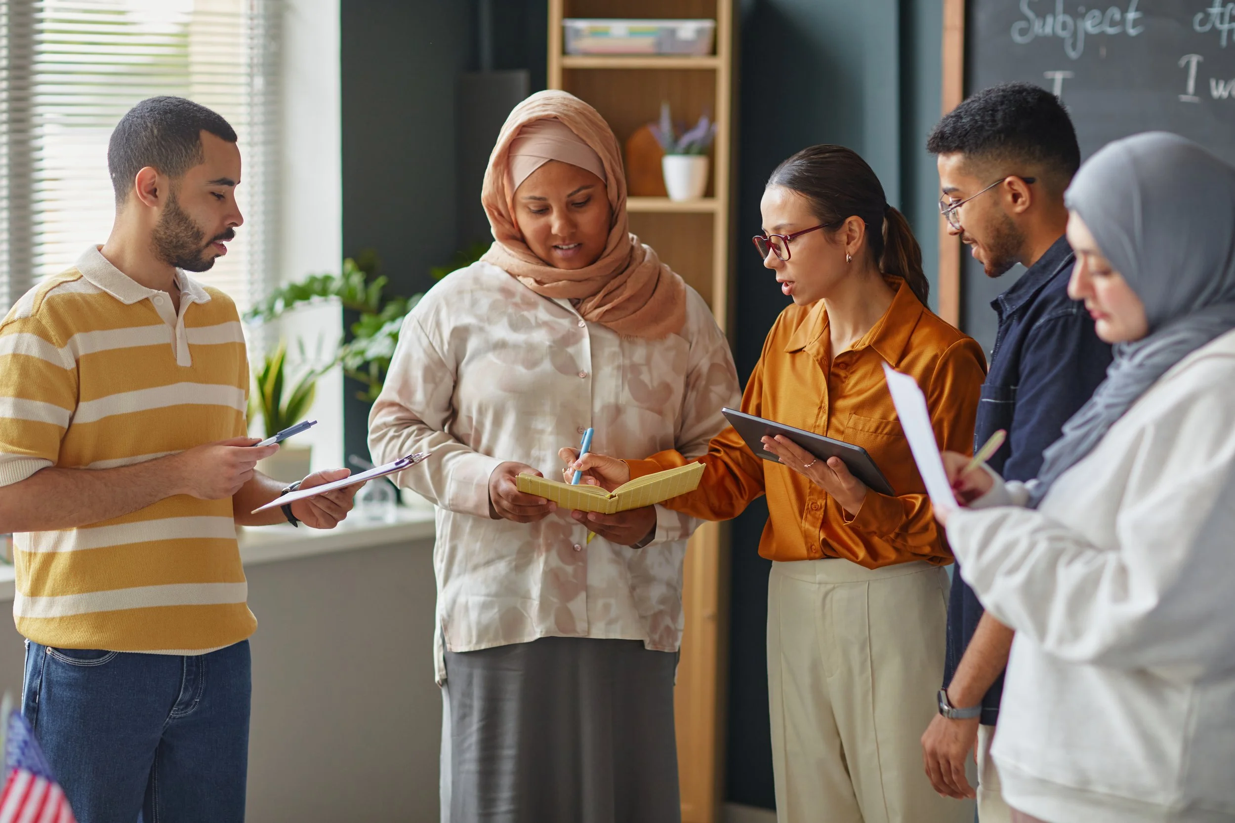 Group of six diverse adults in a classroom discussing, with some holding notebooks and pens, and a blackboard in the background.