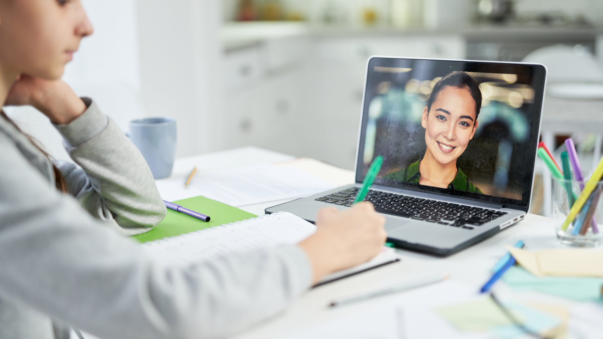 Person on a video call with a woman smiling on the laptop screen, sitting at a desk with papers and colorful pens.