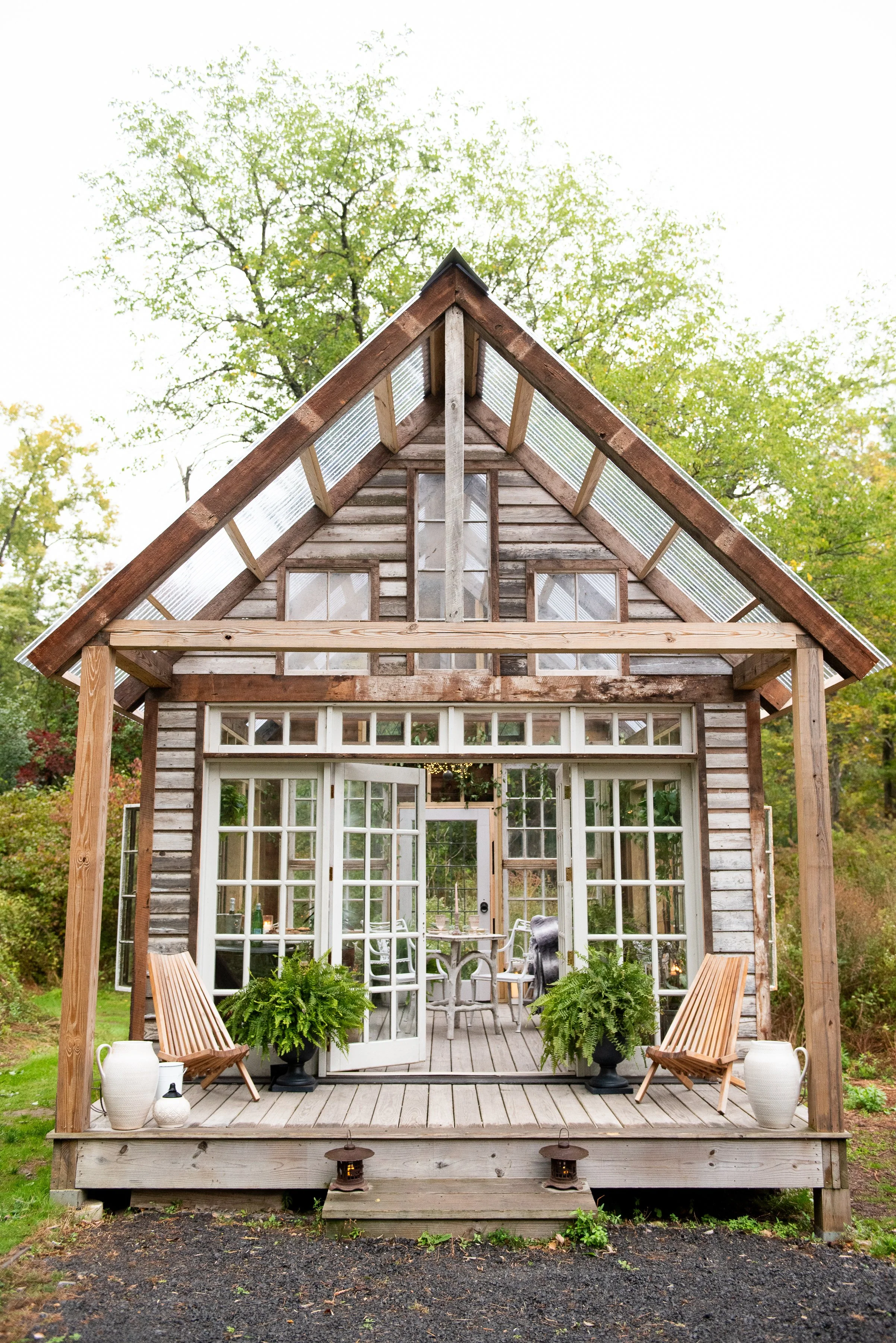 A wooden house with large glass doors and windows, surrounded by greenery. The front porch has two wooden chairs, potted plants, and decorative lanterns, with a ladder leading up to the porch from a gravel area.