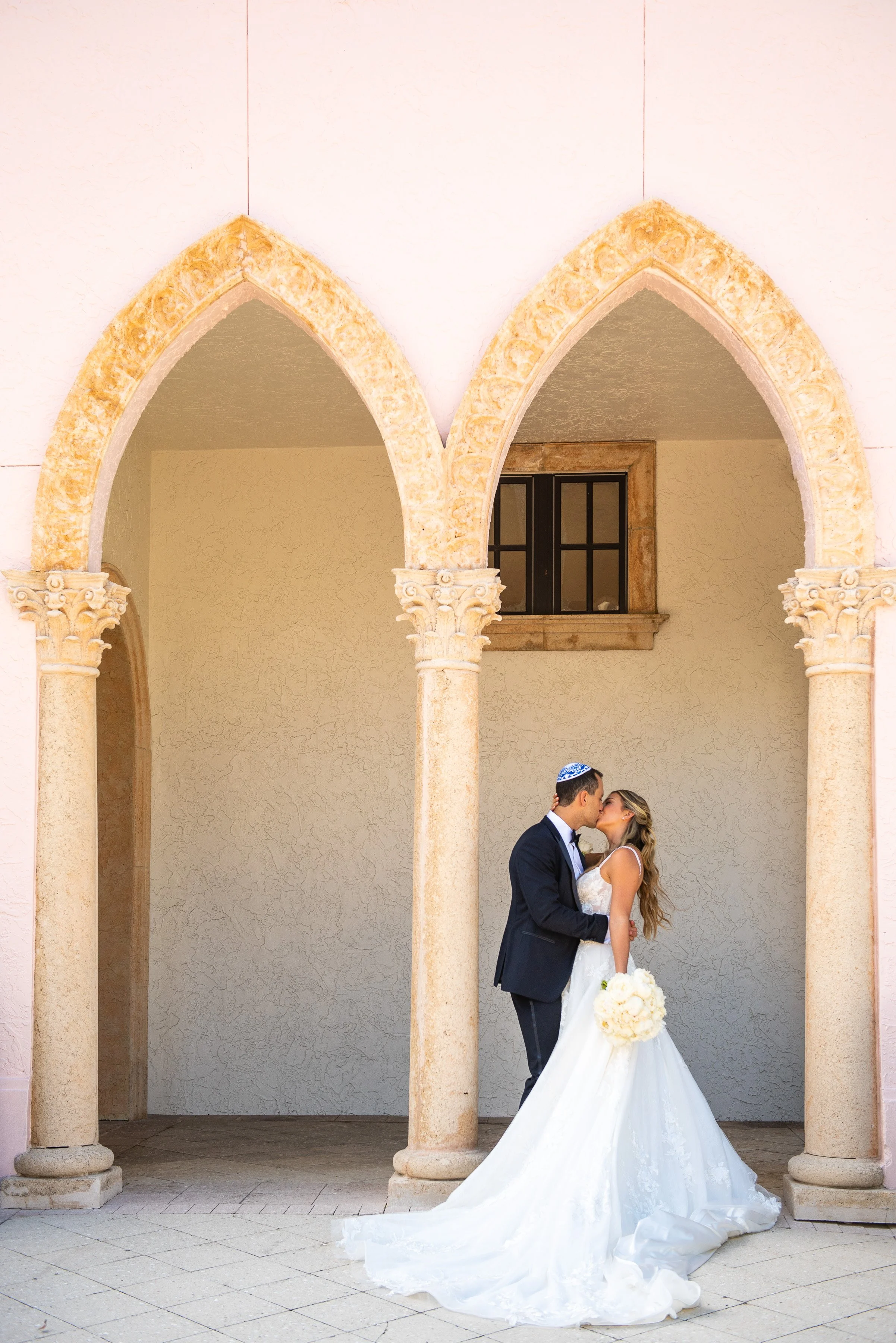 A bride and groom sharing a kiss underneath stone arches at a wedding venue. The bride is holding a bouquet, and the groom is wearing a dark suit with a white shirt and a blue kippah.