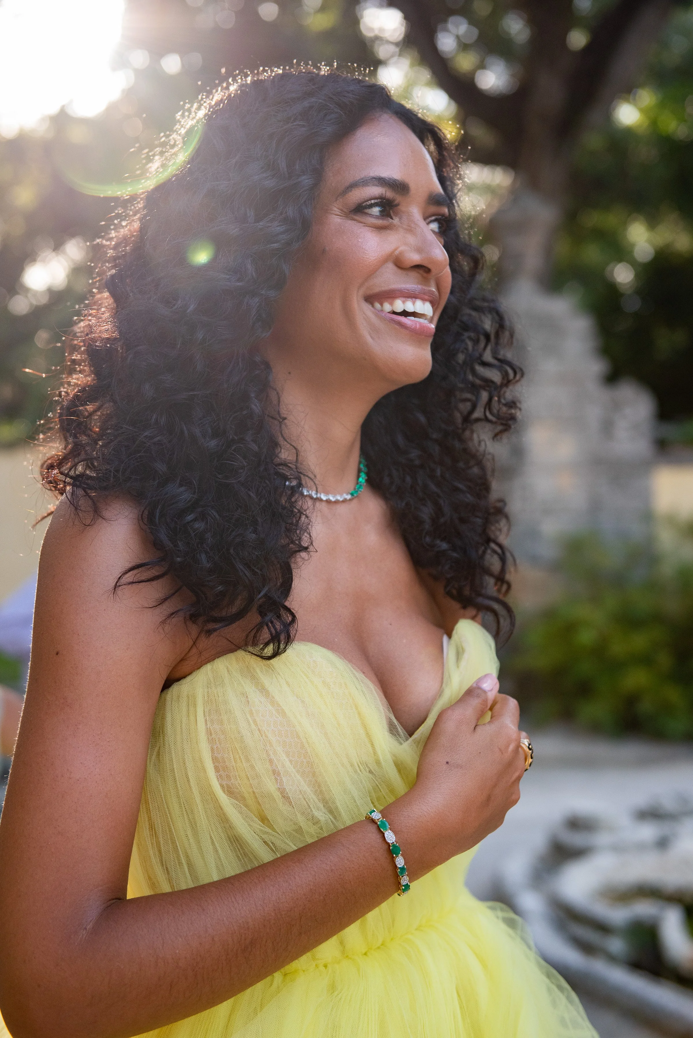 A woman with curly dark hair and medium skin, smiling and looking to the right side in an outdoor setting with sunlight and blurred greenery in the background. She is wearing a yellow strapless dress, a turquoise necklace, a bracelet, and a ring.