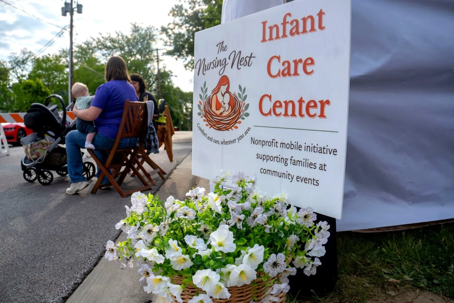 The Nursing Nest Infant Care Center sign with flowers below in a basket. Two women sitting to the left with babies and a stroller