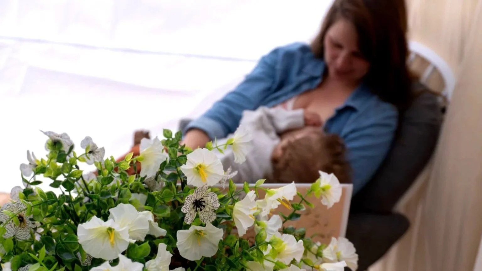 White and green flowers in forefront. Woman breastfeeding her baby in the background on a white and gray rocking chair.