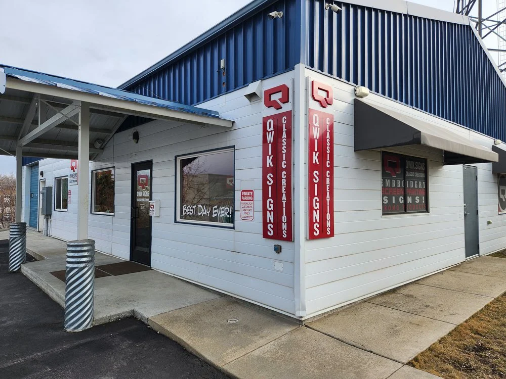 Exterior view of Qwik Signs & Classic Creations in Helena, MT. There's a sidewalk, a few parking space barriers, and a window with a sign that says "Best Day Ever!"