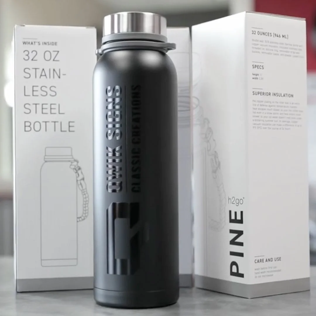 A promotional stainless steel water bottle with a matte silver finish, featuring laser-cut text that says "QWIK SIGNS CLASSIC CREATIONS" in glossy black, placed between two white boxes with product information.