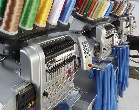 Industrial embroidery machines with colorful spools of thread on top and blue fabrics underneath.