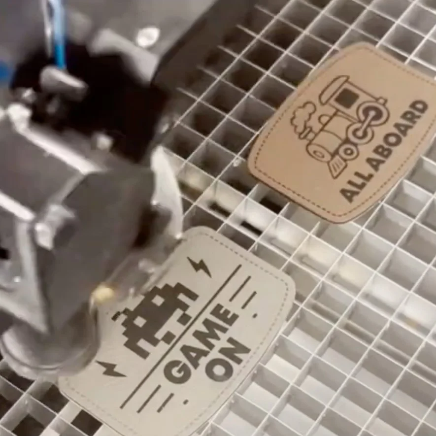 A laser engraving machine printing two designs. The first item has a pixelated video game character with the text "GAME ON". The second item has a drawing of a train with the text "ALL ABOARD".