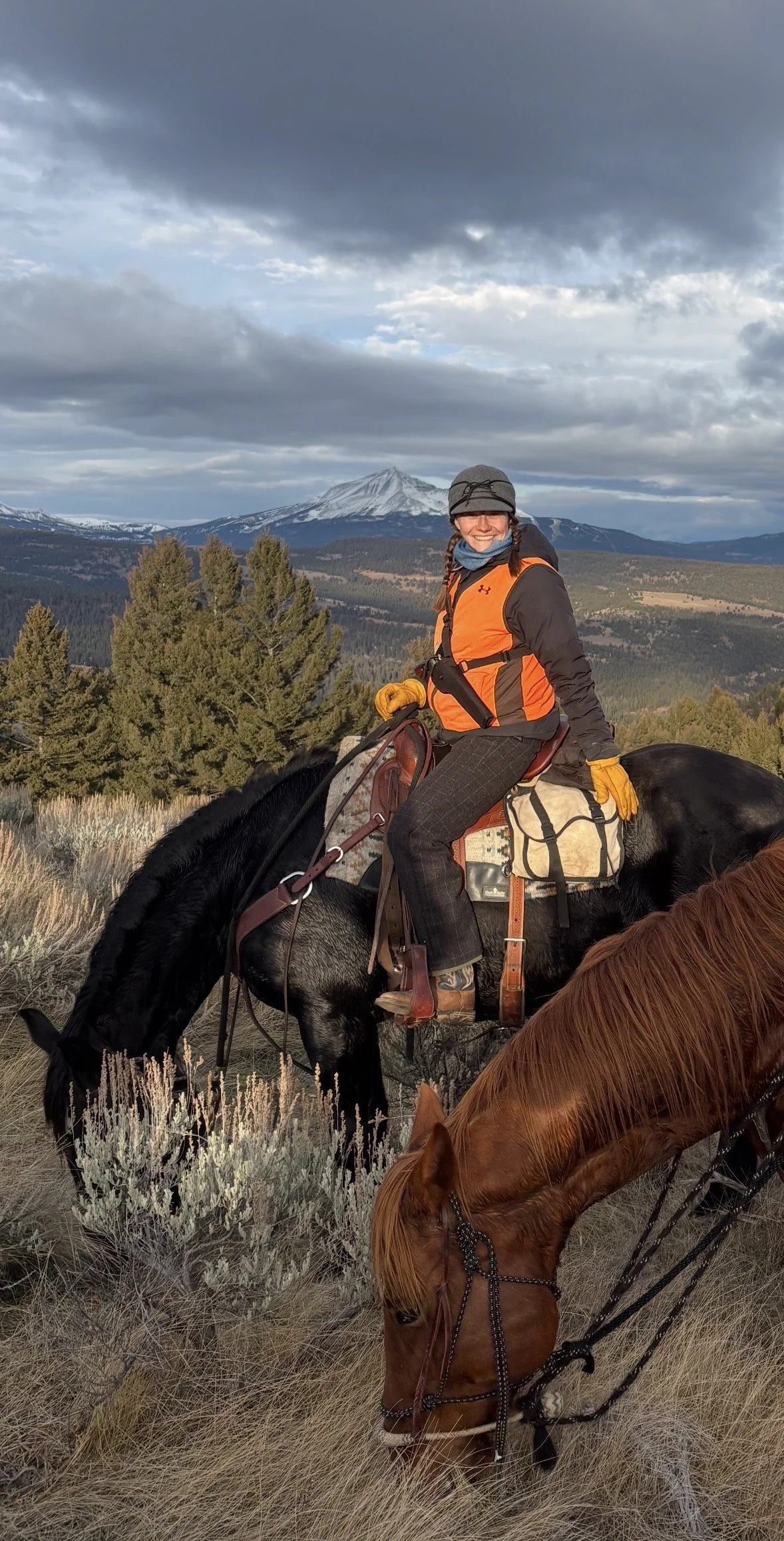 A woman smiling while riding a black horse in a mountainous landscape with snow-capped peaks, trees, and a cloudy sky.