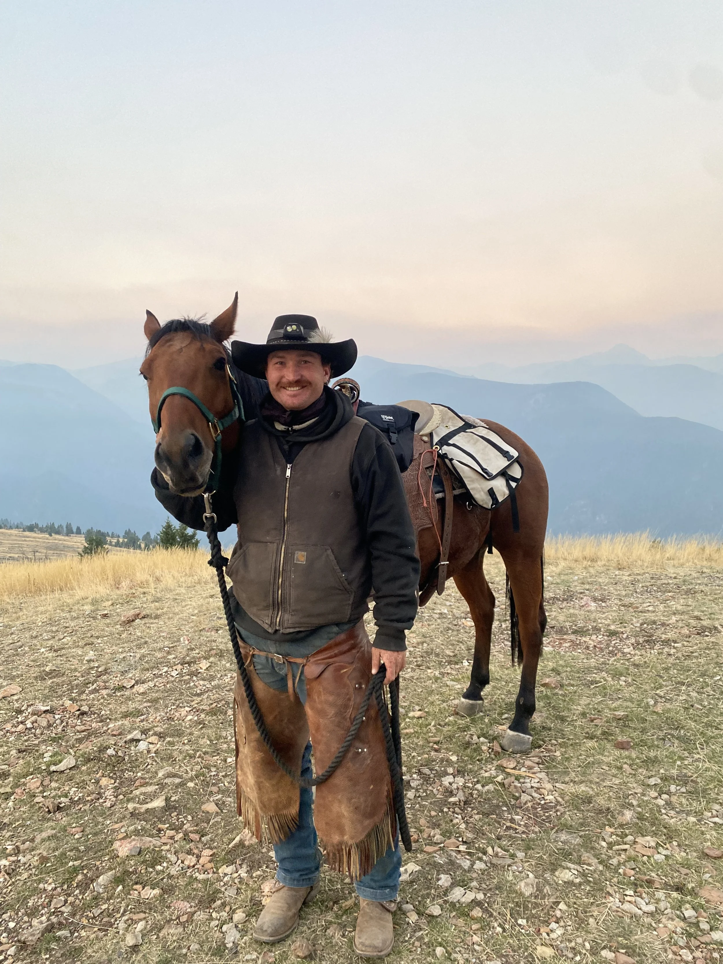 Man dressed in western attire standing outdoors with a horse on a mountain landscape during sunset or sunrise.