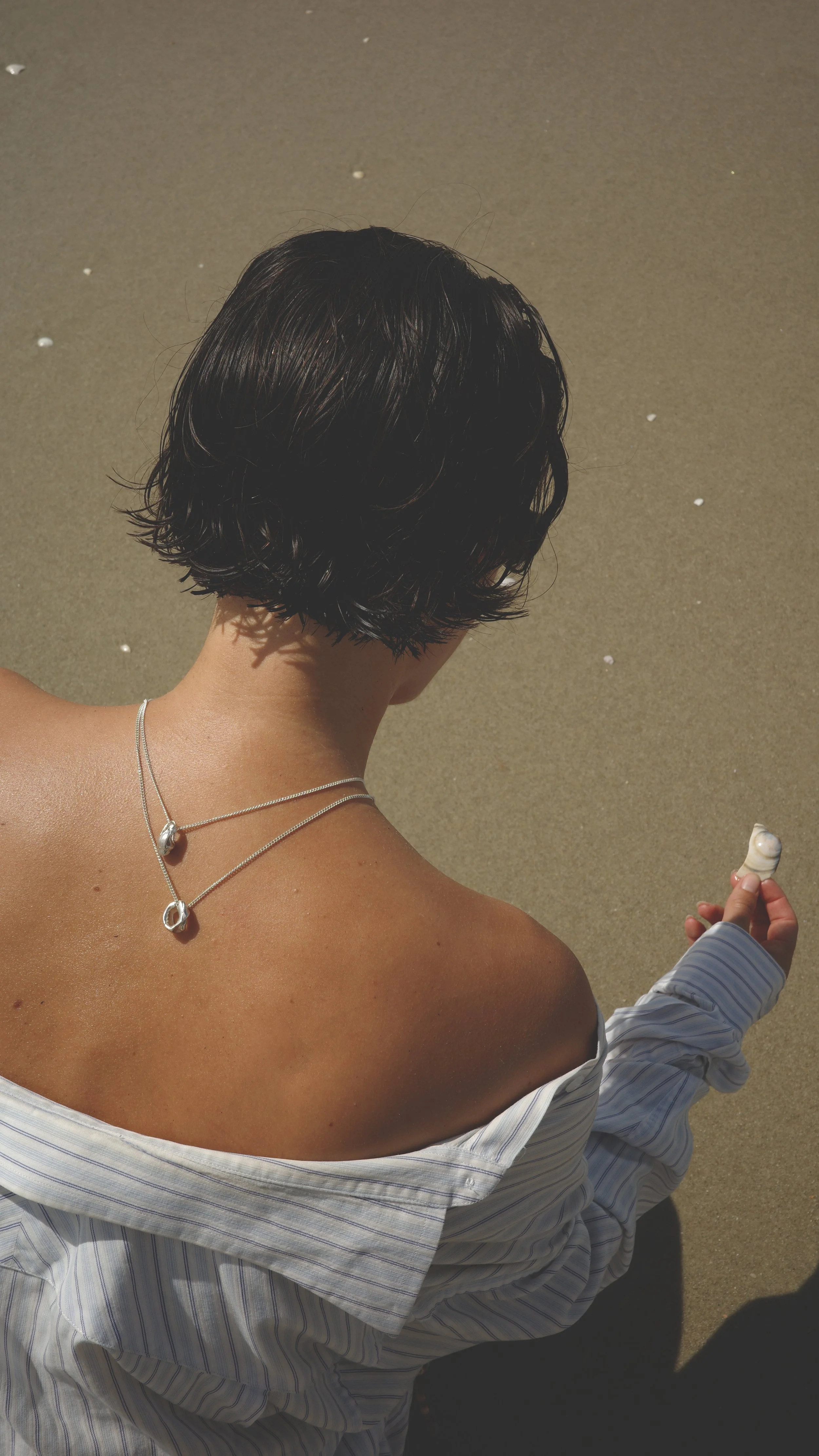 A person with short black hair sitting on the beach, wearing a striped shirt off one shoulder and silver jewelry, holding a seashell.