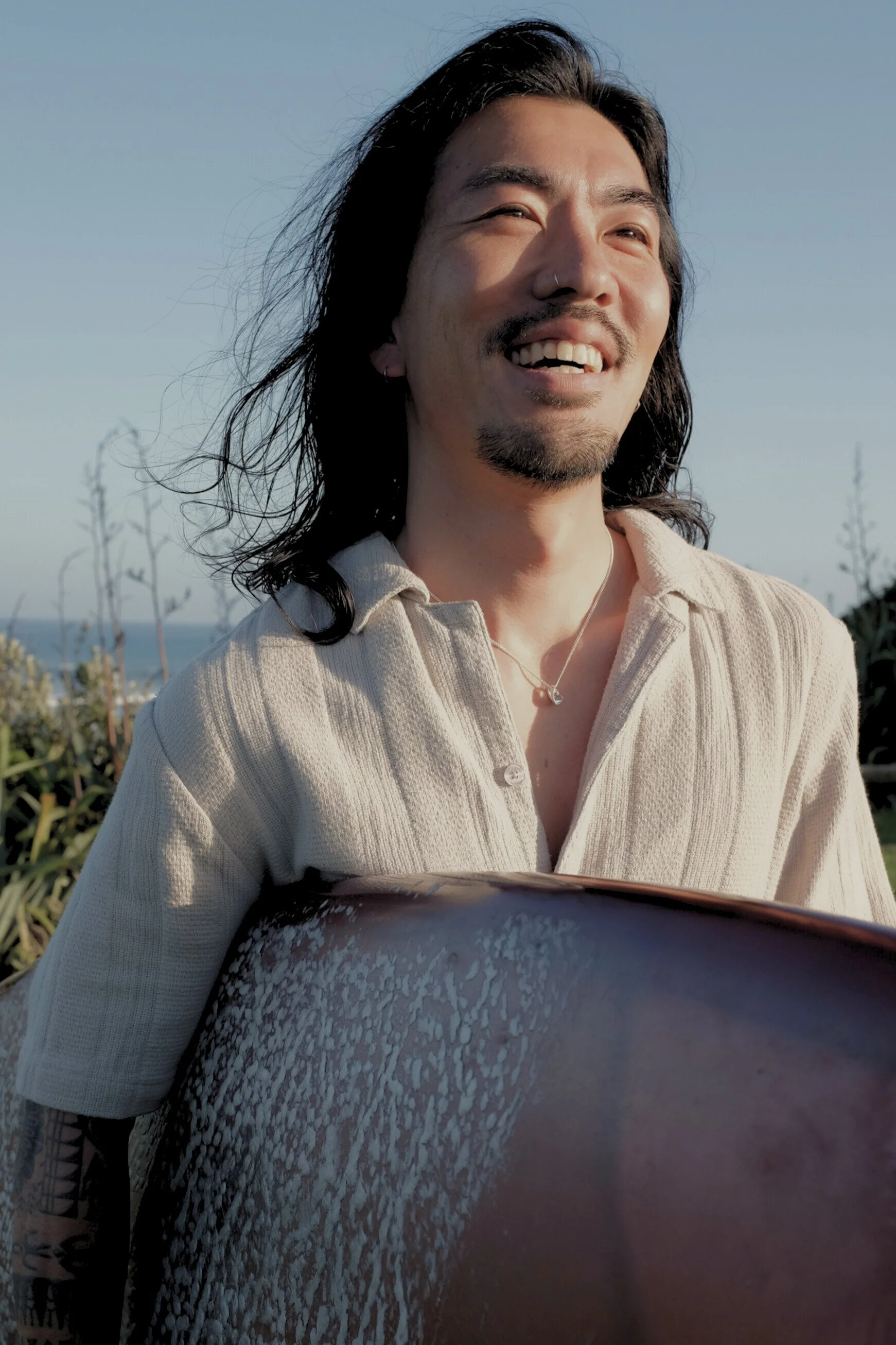 A smiling man with long dark hair, wearing a light-colored shirt and holding a surfboard, standing outdoors under a clear sky.