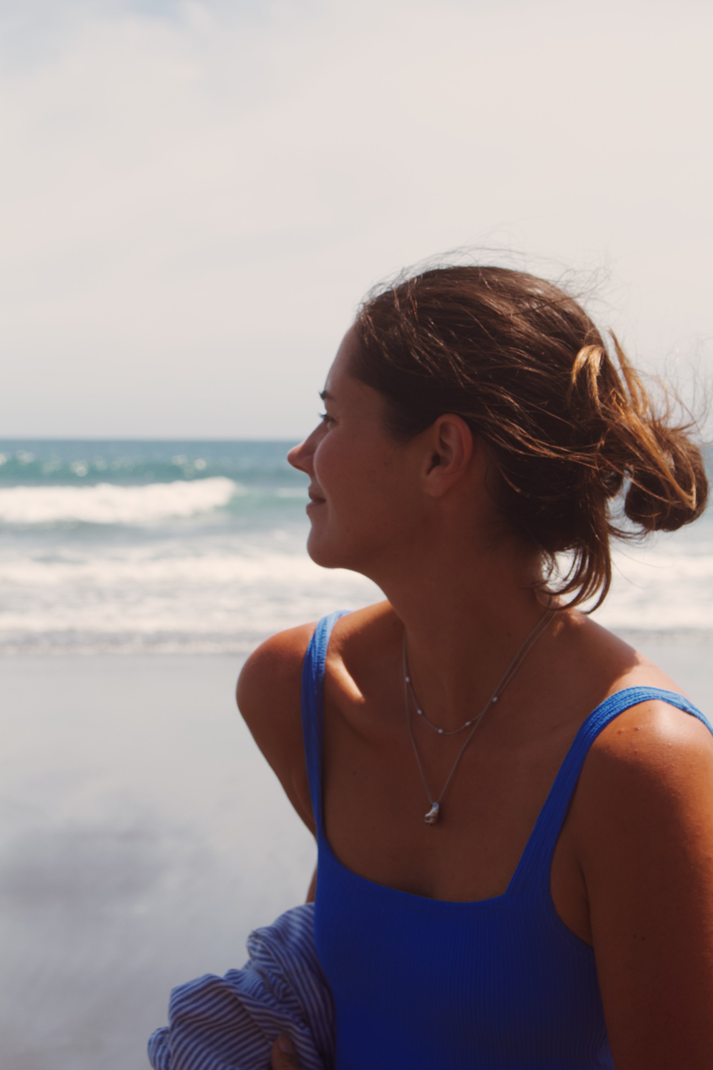 Side profile of a woman with brown hair tied back, wearing a blue sleeveless top, standing on the beach with ocean waves in the background.