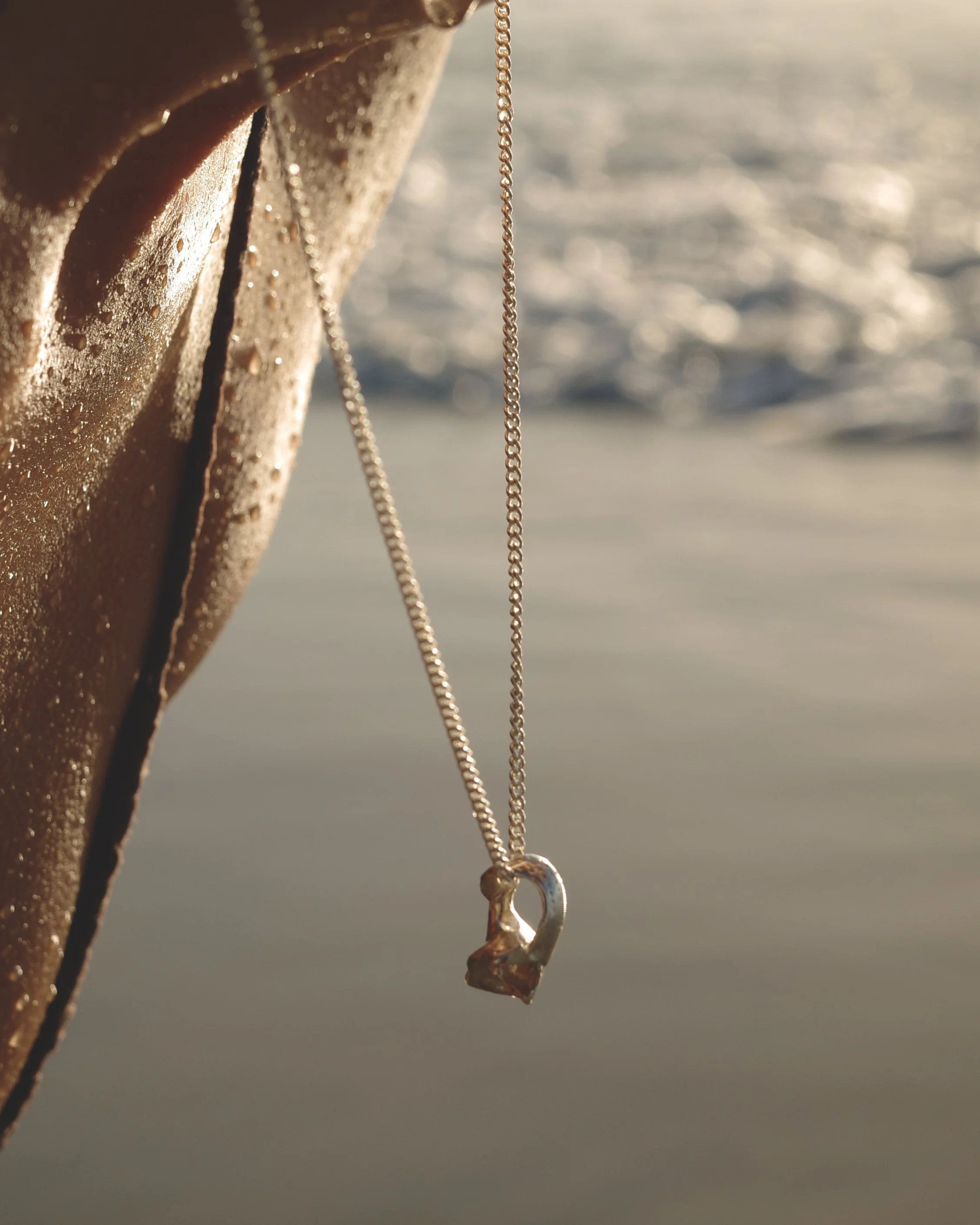 Silver necklace with a shell pendant hanging from a wet, sunlit surface beside water.