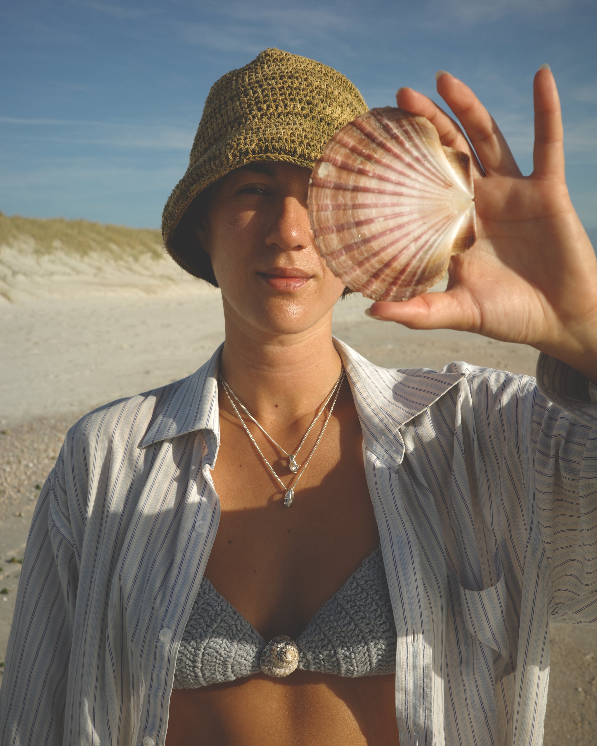 A woman on a beach is holding a scallop shell near her face. She is wearing a brown knit hat, a striped shirt, layered necklaces, and a gray bikini top with a seashell decoration. The beach and sky are visible in the background.
