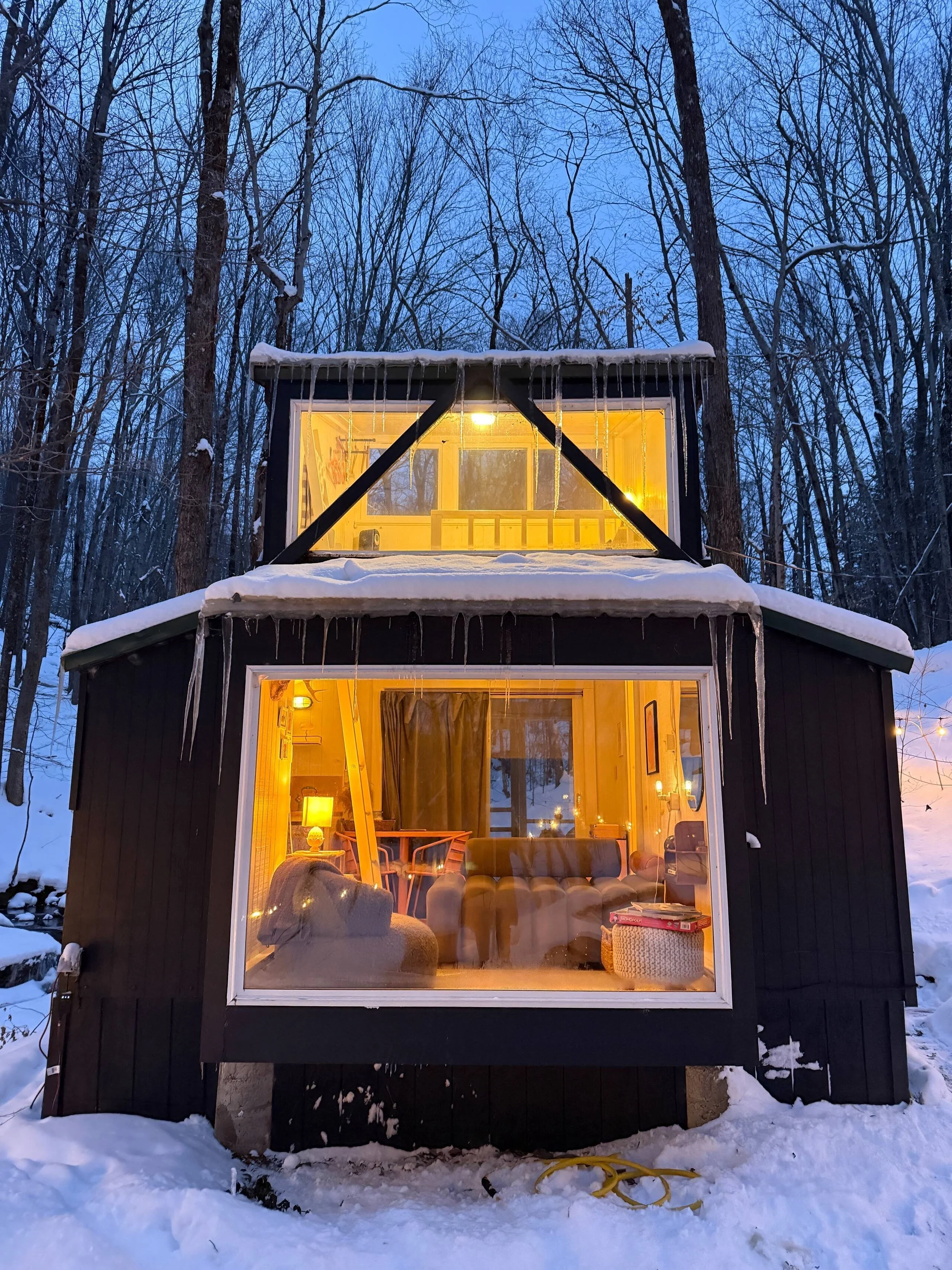A cozy two-story black cabin with large front windows, snowy surroundings, and icicles hanging from the roof, illuminated warmly from inside in a winter forest setting.