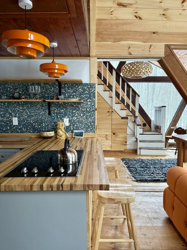 Interior view of a cozy wooden house with a kitchen island, orange pendant lights, wooden staircase, and a patterned rug.