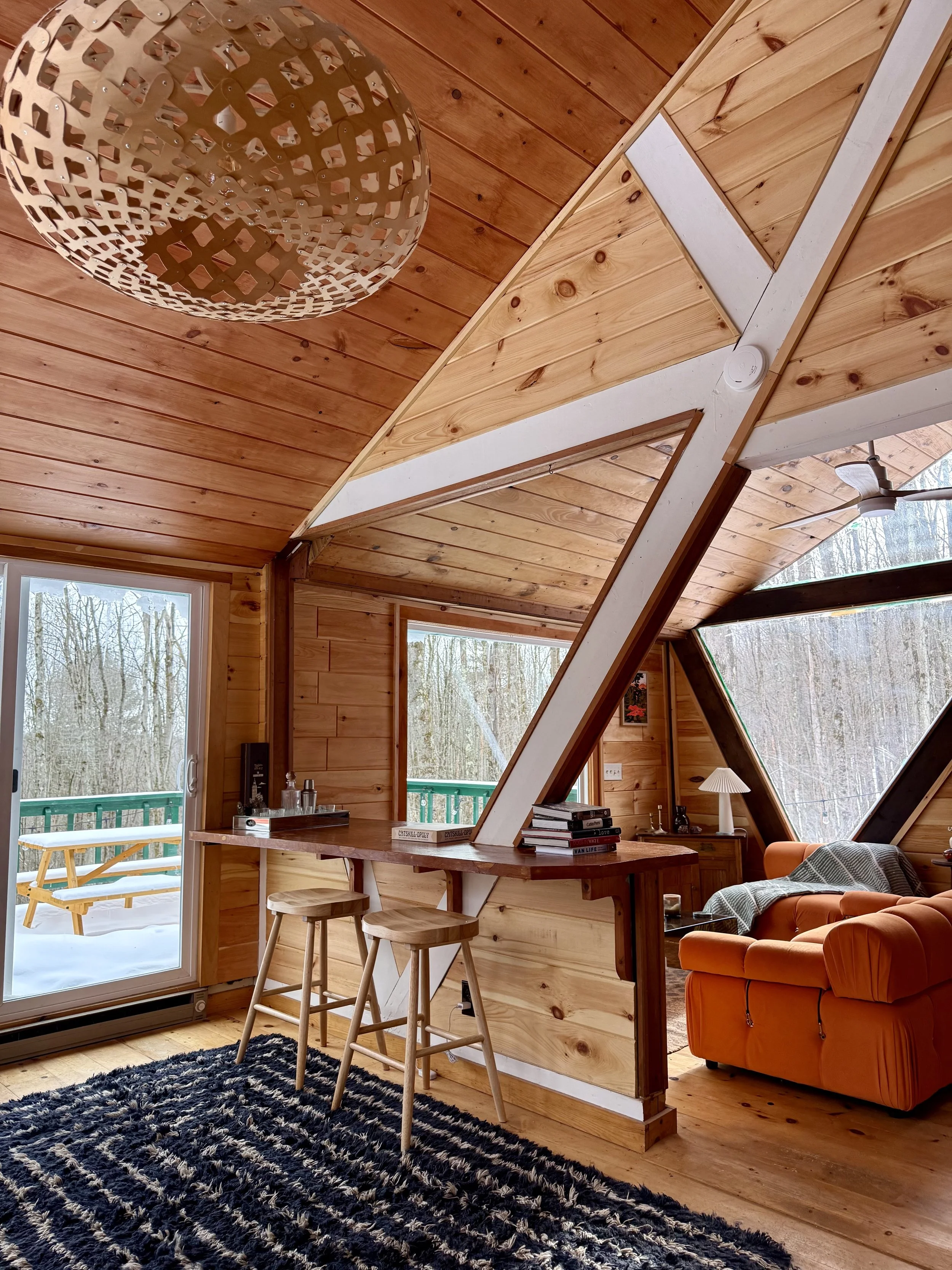 Interior view of a cozy wooden loft with large windows, a black and white striped rug, two bar stools at a small breakfast bar, an orange couch with a throw blanket, and an outdoor scene with snow visible through the windows.