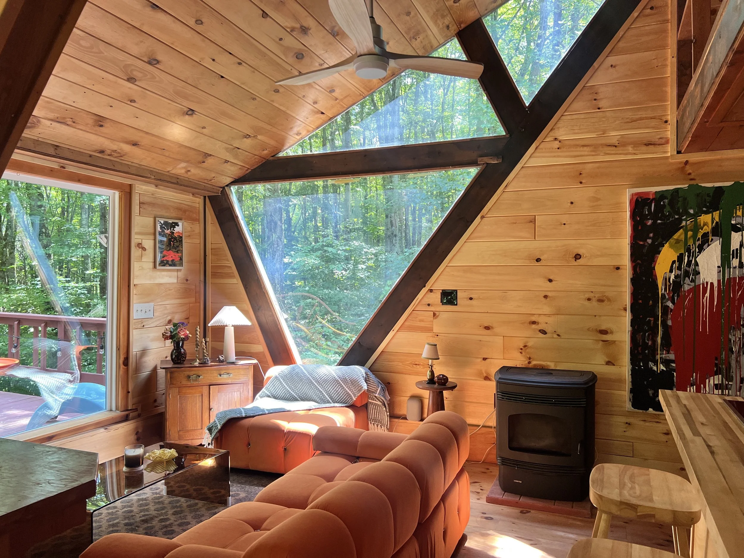 Cozy living room in a wooden cabin with large triangular windows showing a lush green forest outside. The room has a peach-colored sofa, a small wooden side table with a lamp and a potted plant, a wooden cabinet, a modern small black stove, and colorful abstract art on the wall.