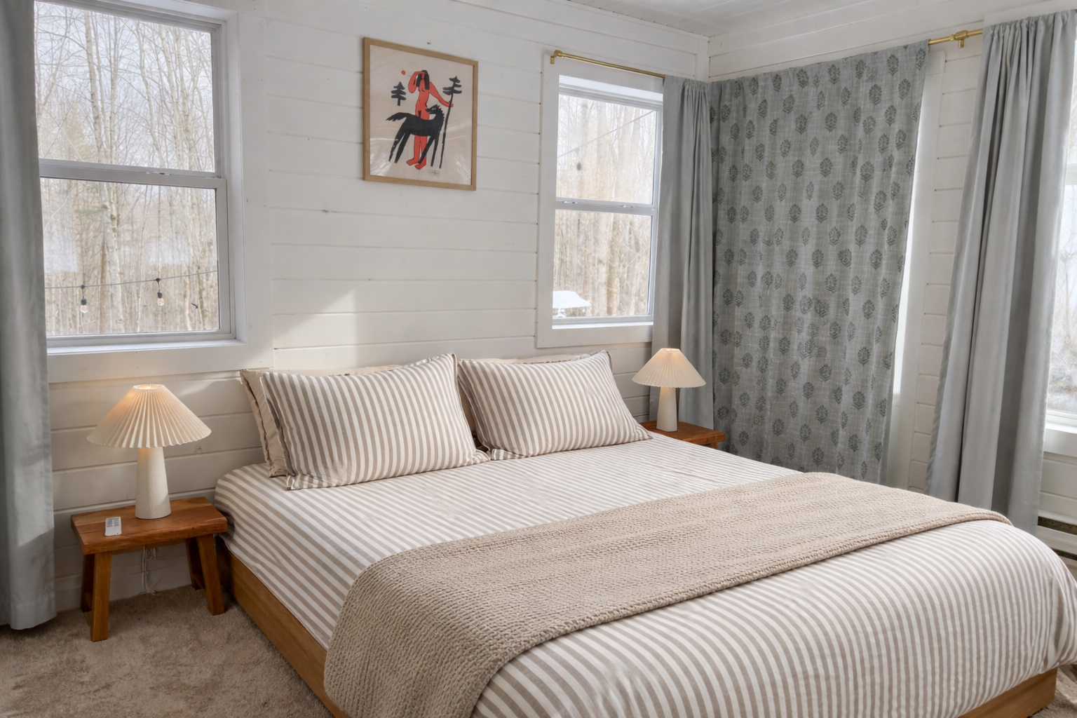 A bedroom with a bed covered with beige and white striped bedding and matching pillows. Two small wooden nightstands with lamps are placed on either side of the bed. The room has two windows with curtains, one with a light blue curtain and the other with a patterned curtain. A framed artwork depicting a stylized animal is on the wall above the bed.
