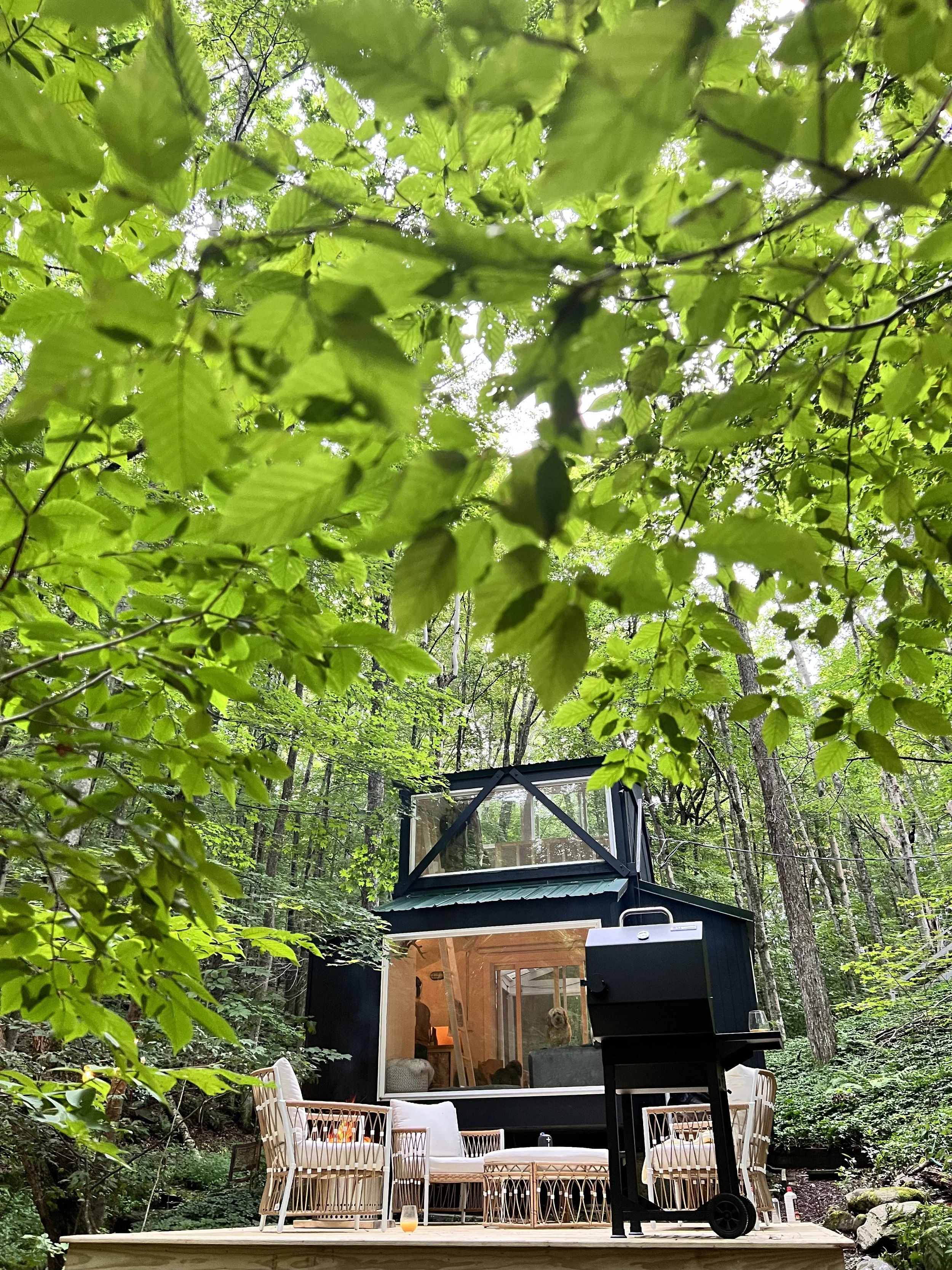 A cozy tiny house with large windows is situated in a lush green forest, featuring a sitting area with four chairs and a small table on a wooden deck, with a black grill nearby.