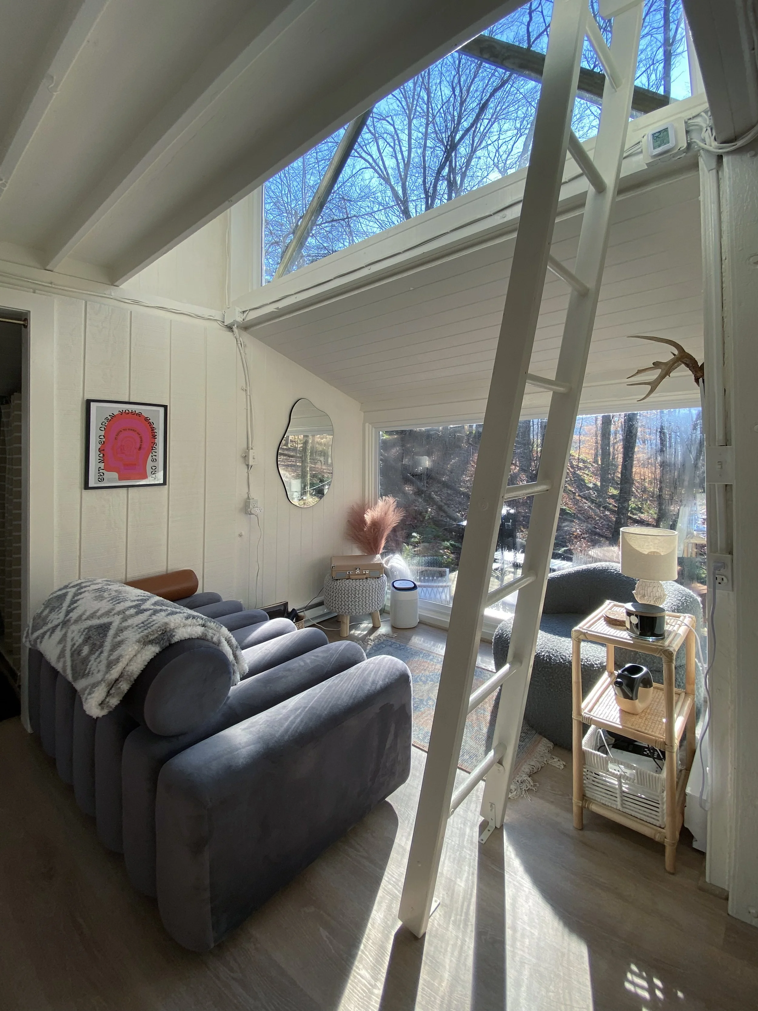 A cozy living room with a loft area, featuring a large window that shows trees outside. There is a gray couch with a blanket, a small wicker table with pink feathers, a mirror on the wall, and a side table with a lamp and other items. A white ladder leads up to the loft space.