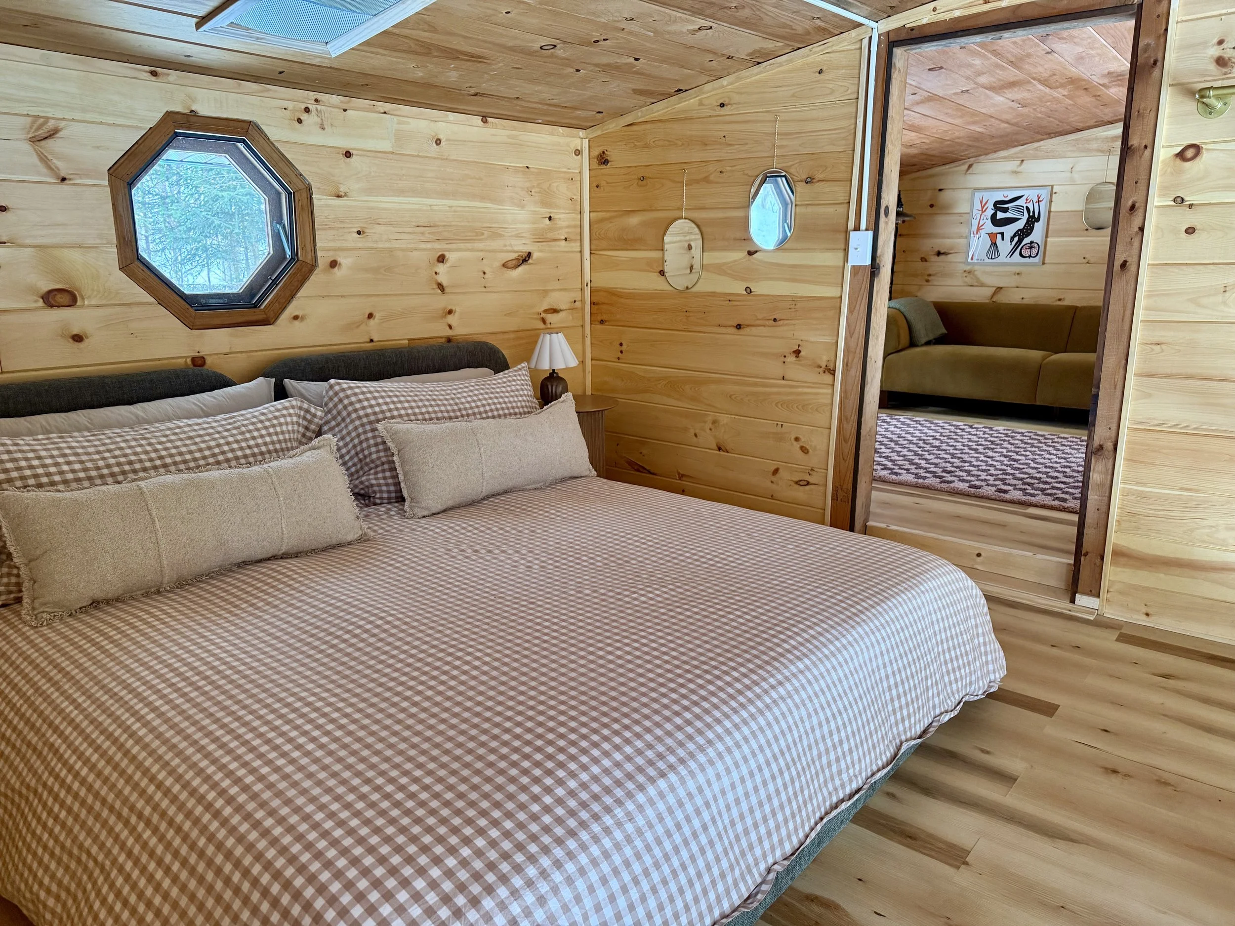 A cozy bedroom with wood-paneled walls and ceiling, featuring a bed with brown and white gingham bedding and multiple pillows, a nightstand with a lamp, and octagonal windows. An open doorway leads to a living room with a green sofa and wall art.