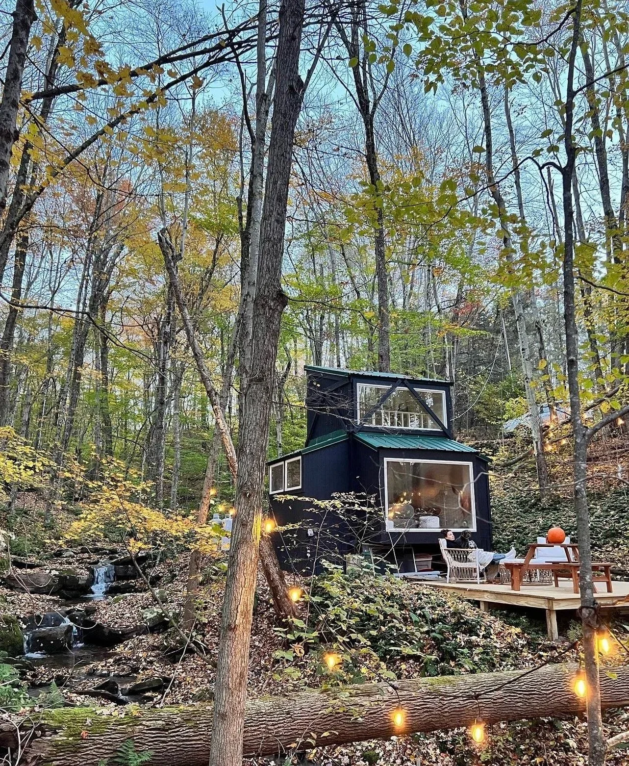 A tiny modern black cabin with large windows is situated in a wooded area with fall foliage. String lights and a small waterfall stream are visible, with a person sitting on a porch.