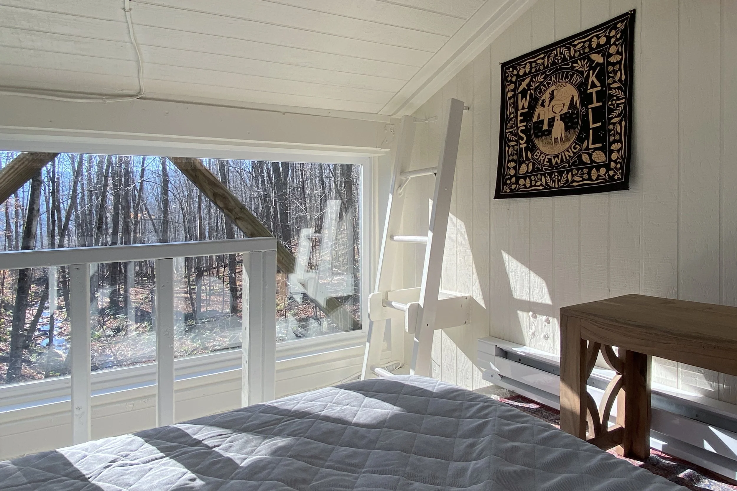 A bedroom with a bed, a white ladder chair, a wooden table, and a wall hanging with a brewing company logo next to a large window showing a wooded outdoor scene.