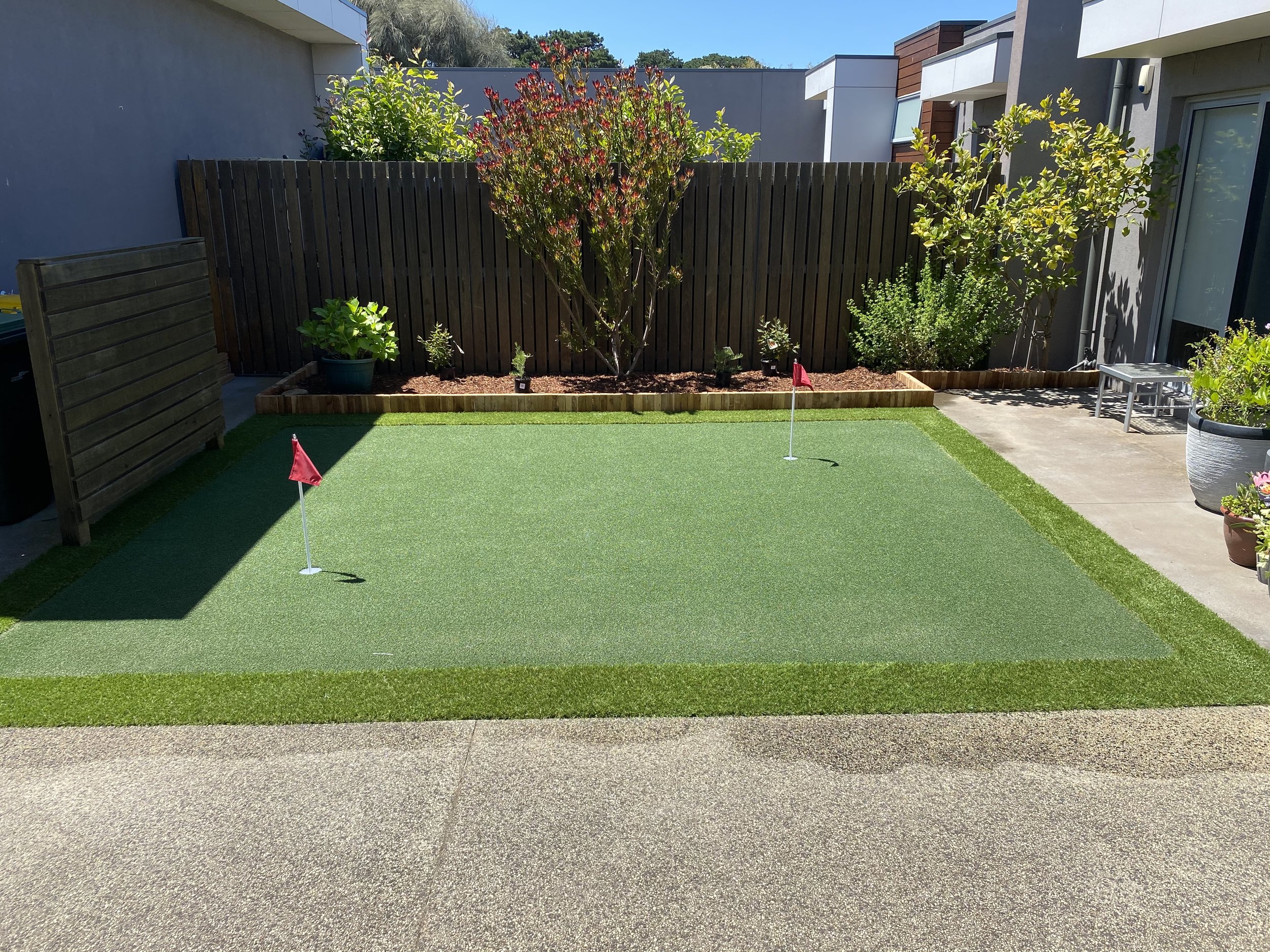 Backyard with artificial putting green, two small red flags, and landscaped garden area with trees and shrubs behind a wooden fence.