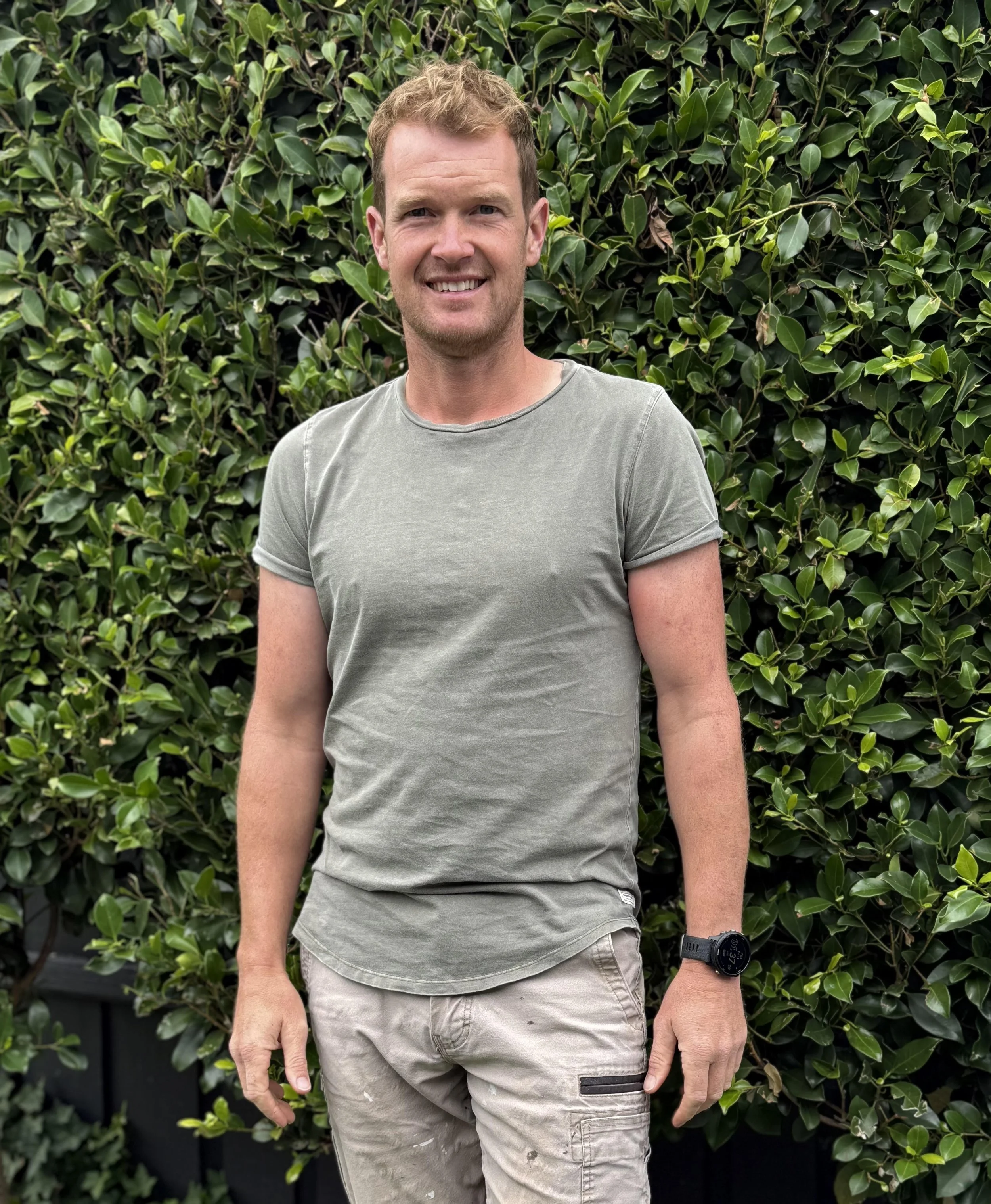 A man smiling while standing outdoors in front of a green hedge, wearing a light gray t-shirt, beige cargo pants, and a black watch.