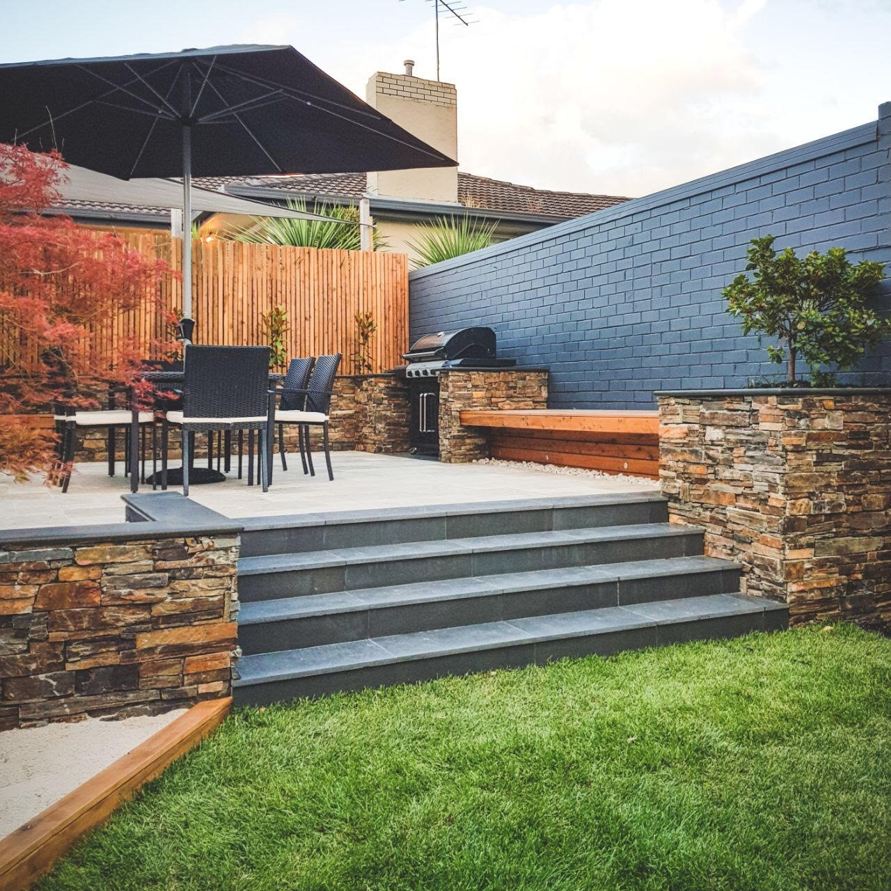 A backyard patio with dark gray steps leading to a tiled outdoor space. There is a round table with four chairs and a large black umbrella. A built-in outdoor grill is in the corner, and a wooden bench is attached to a blue brick wall. The area is enclosed by a wooden and stone fence, with some greenery, including a small shrub and trees.