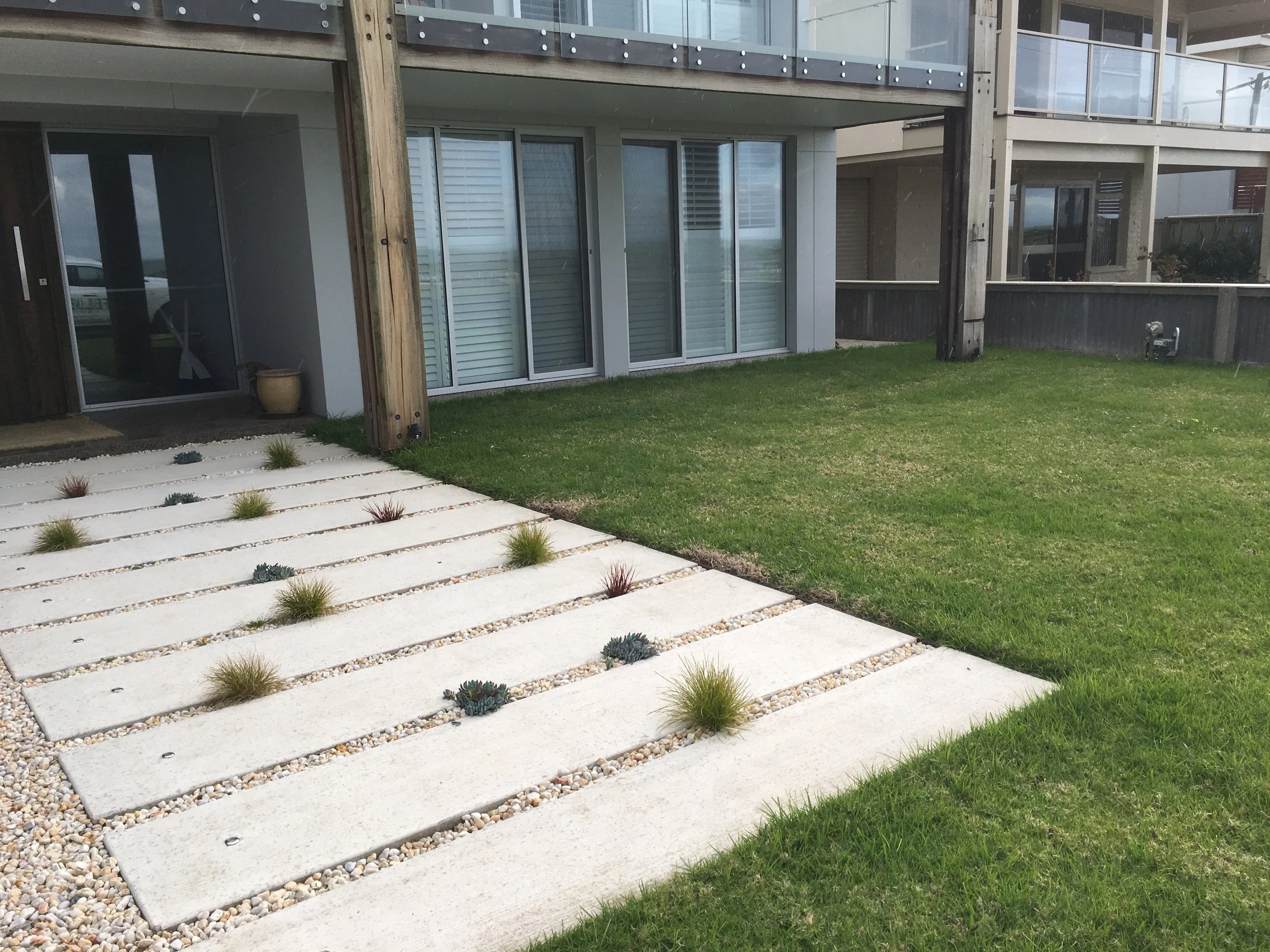 Modern patio and lawn with concrete slabs and small desert plants outside an apartment building with glass balconies.